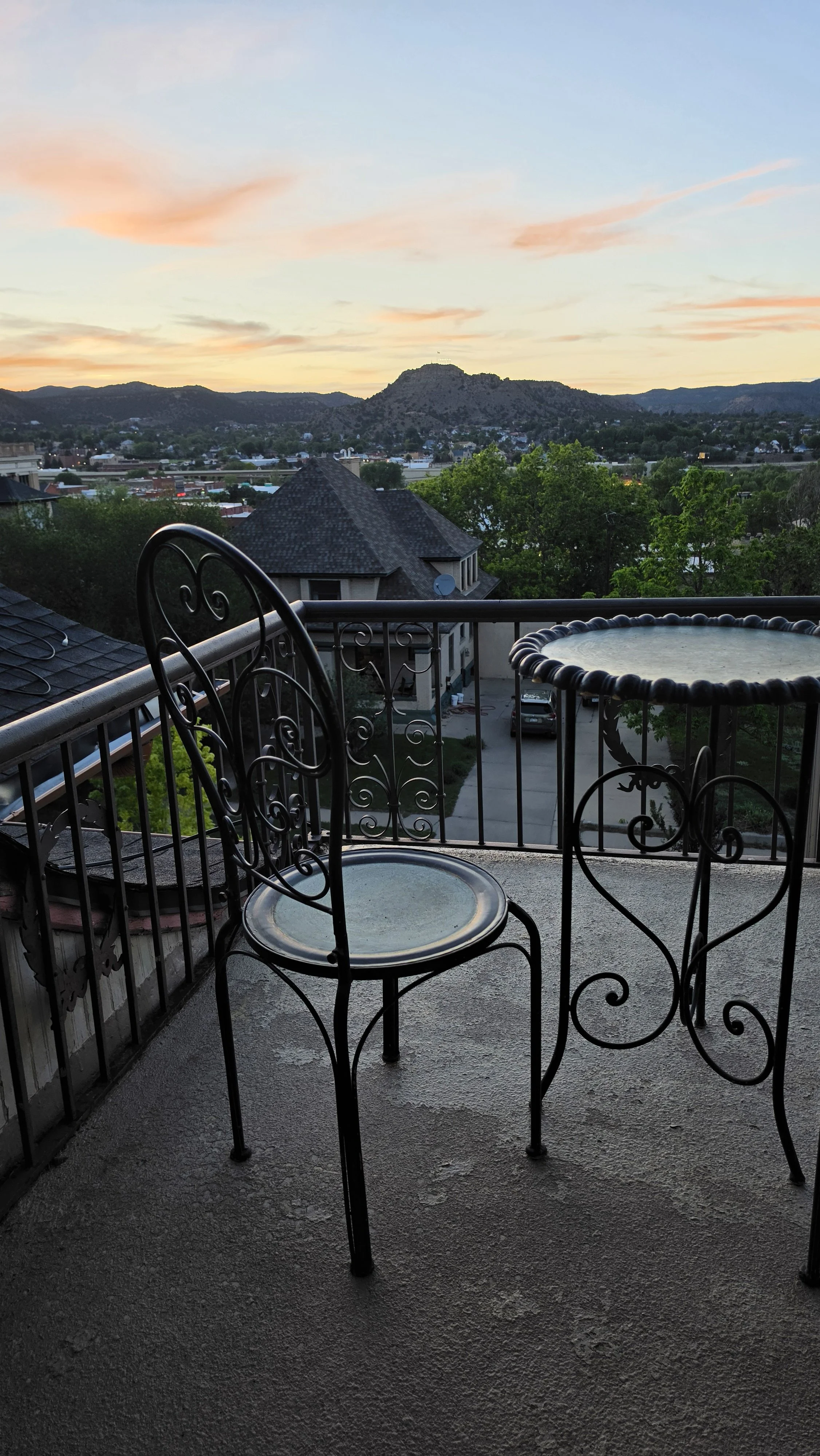 View of a backyard balcony with wrought iron chair and table, overlooking a suburban area with houses, trees, and mountains in the distance at sunset.