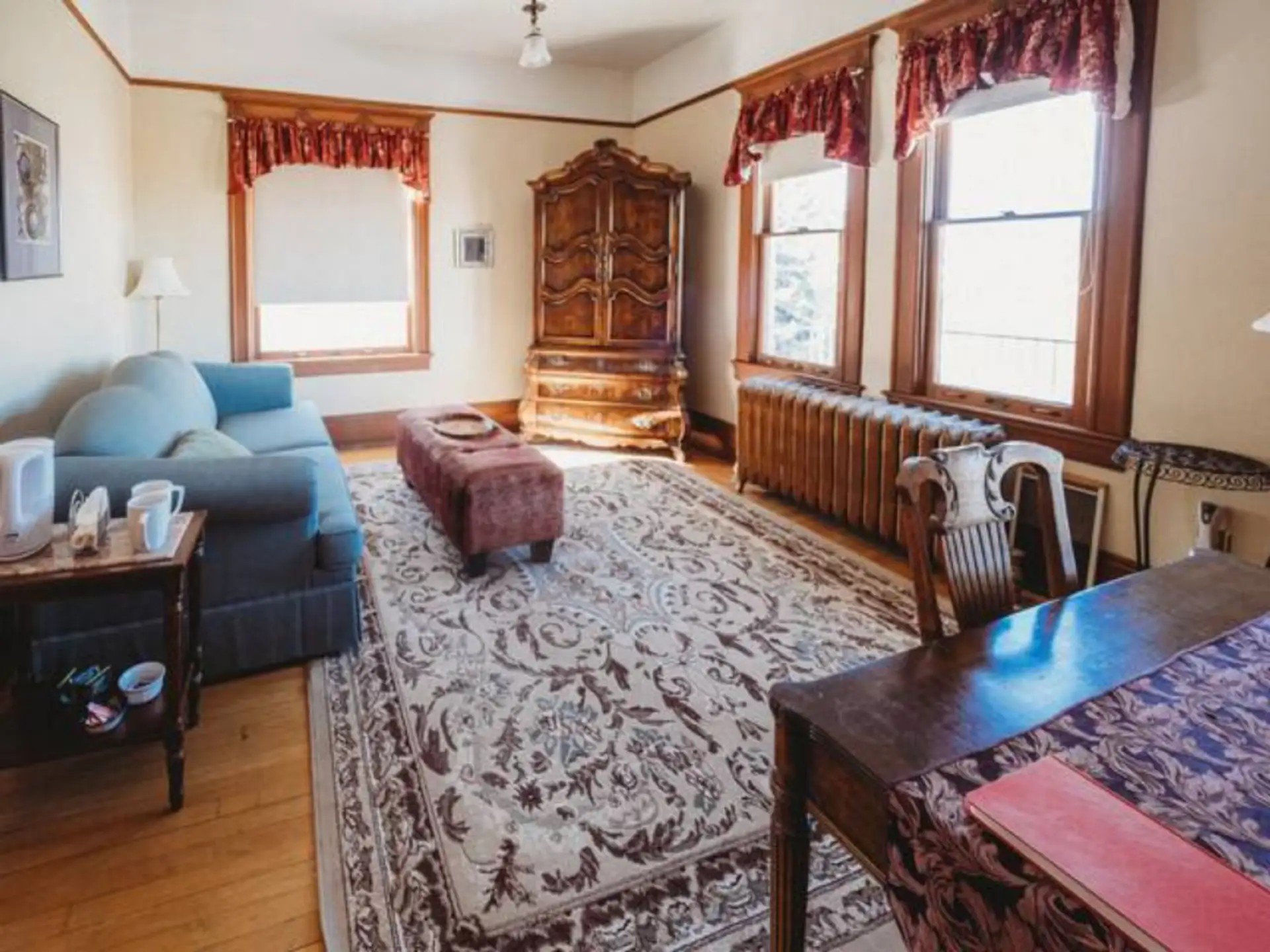 Living room with wooden framed windows, a patterned rug, a light blue sofa, wooden furniture including a cabinet, and curtains with red valances.