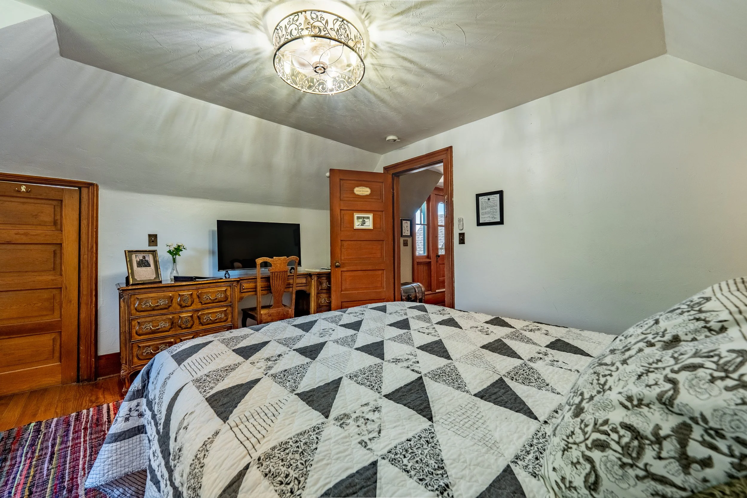 A bedroom corner with a bed featuring a black, white, and gray quilt, a wooden dresser topped with a framed photo and a glass vase with flowers, a flat-screen TV, a wooden chair, two wooden doors, and framed pictures on the walls. A decorative ceiling light casts shadows on the ceiling and walls.