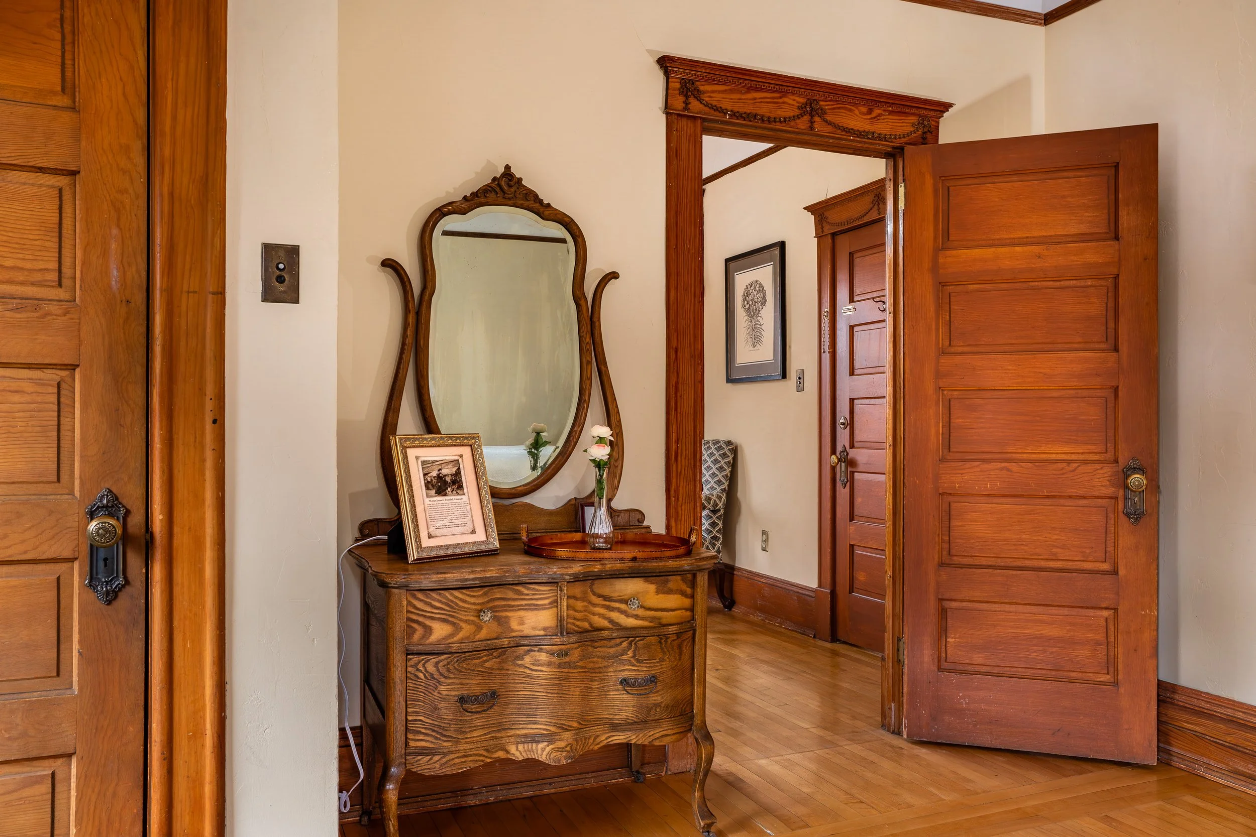 A vintage wooden dresser with framed photographs and decorative items, placed against a wall in a room with wooden trim and floors, wooden doors partially open.
