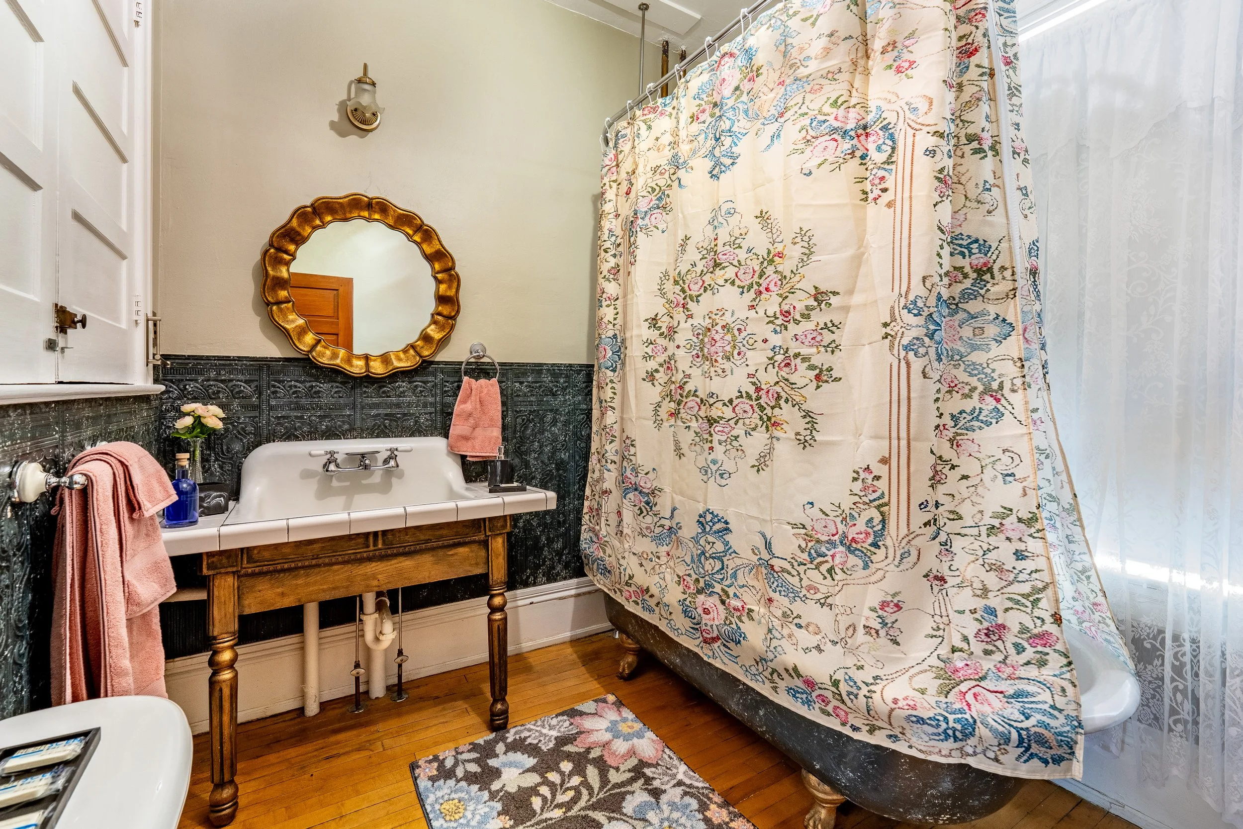 A vintage bathroom with a white porcelain sink, a gold-framed oval mirror, and a floral fabric shower curtain. Pink towels and a small vase of flowers are on the sink, and a wooden table supports the sink beneath. The bathroom has wood flooring, a shaded window, and decorative wall tiles below the plain upper wall.