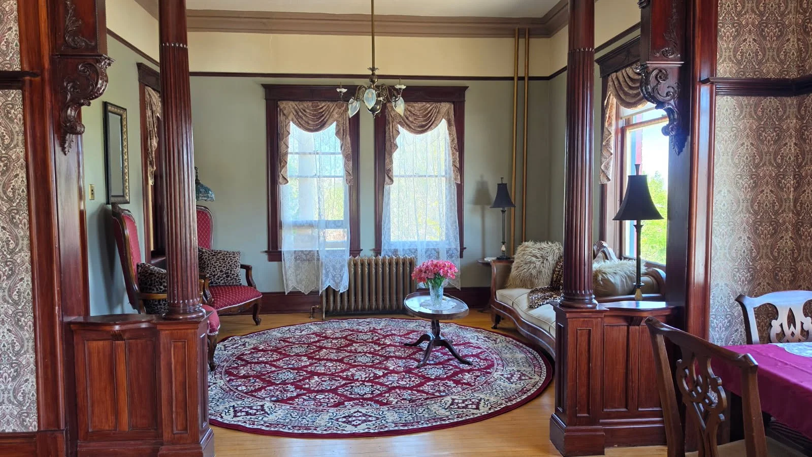 Living room with Victorian-style decor, featuring dark wood trim, patterned curtains, and a round rug with floral design. There are two windows with lace curtains, a small coffee table with pink flowers, and various chairs and lamps.
