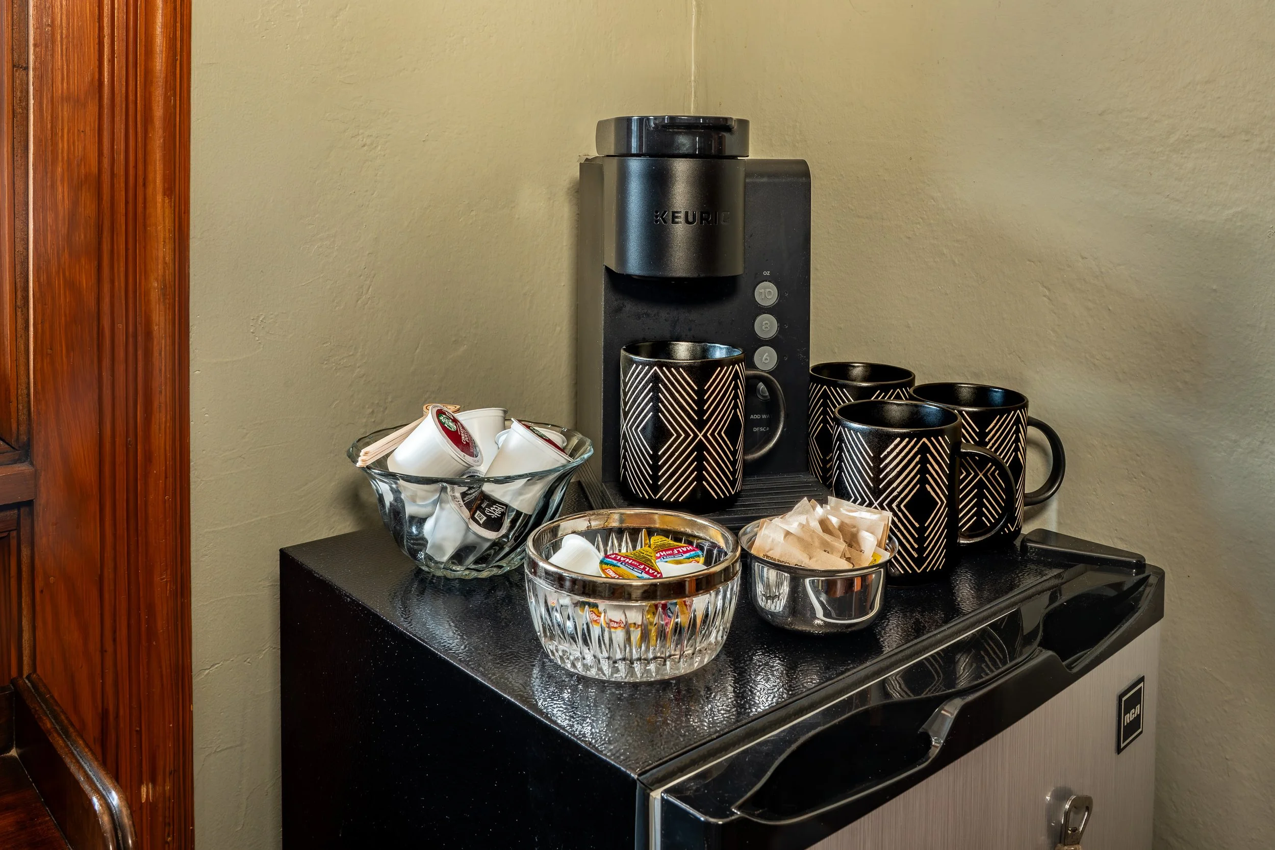 A coffee station with a Keurig machine, black and white patterned mugs, and various coffee condiments and accessories on a black countertop.