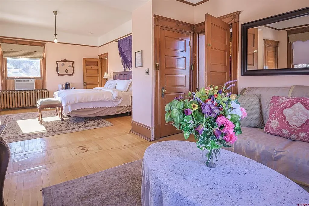 A cozy bedroom with a large bed, wooden furniture, a window with natural light, and a floral arrangement on a covered table.