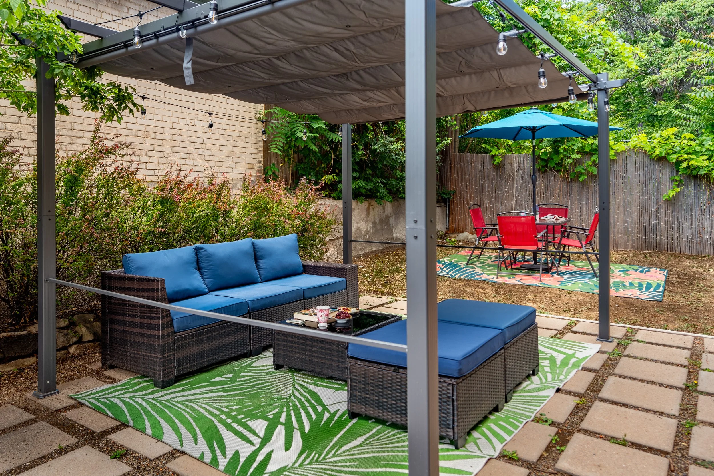 An outdoor patio area with a black wicker sofa with blue cushions under a metal pergola with string lights. There is a small black table with cups on it, a colorful outdoor rug with a green leaf pattern, a circle table with four red chairs, a blue umbrella, and lush green plants and trees in the background.