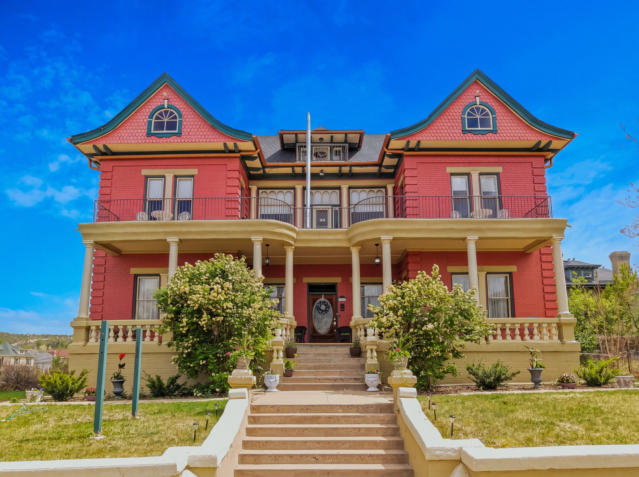 A large, colorful Victorian-style house with a pink and yellow exterior, multiple stories, and decorative elements. The house has a front porch with white columns, stairs leading up to it, and various potted plants and bushes in the garden area. The sky is clear and blue.