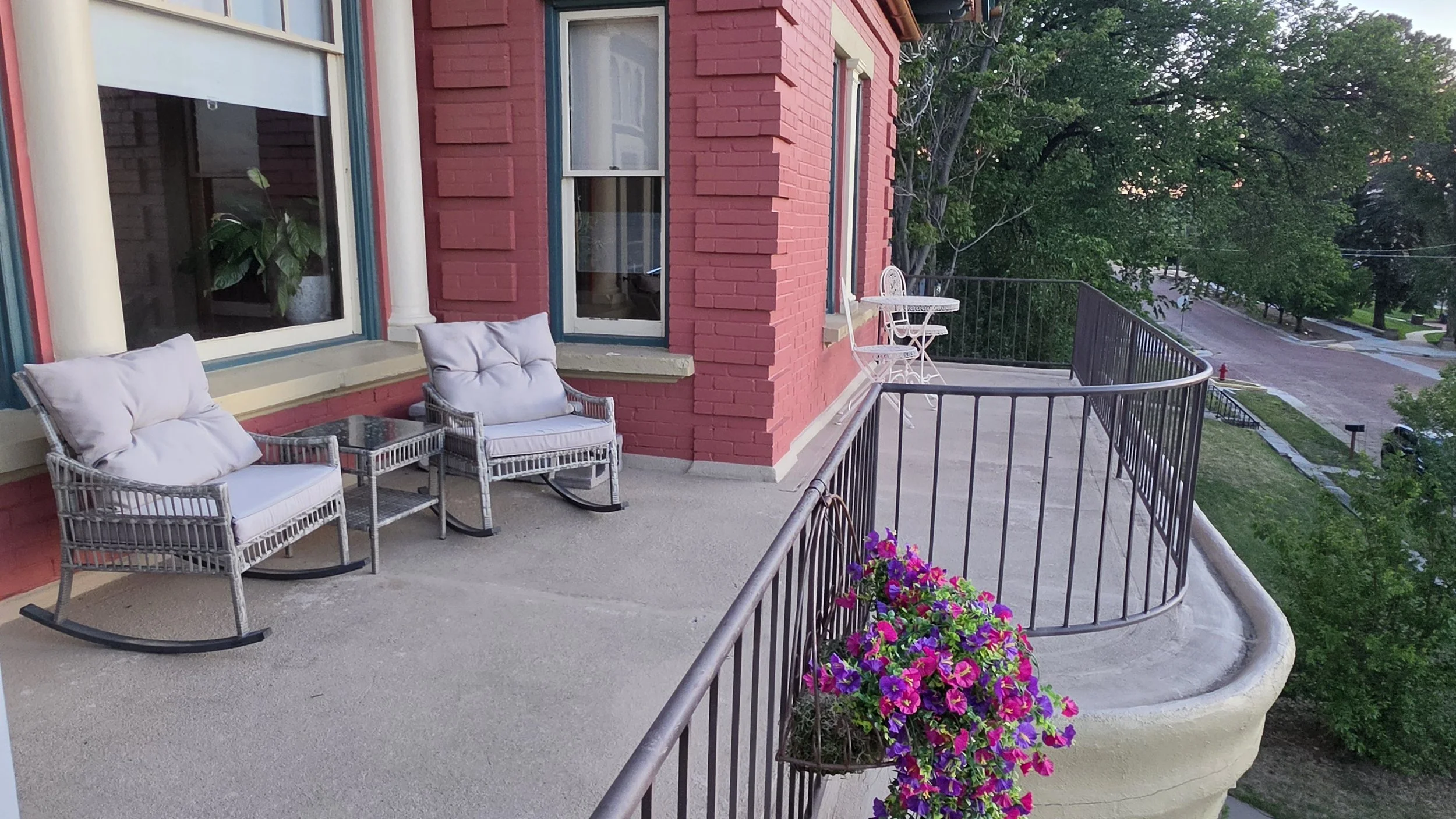 A balcony with two wicker chairs with white cushions, a small table, a flower box with pink and purple flowers, and a view of a neighborhood with trees and a street.