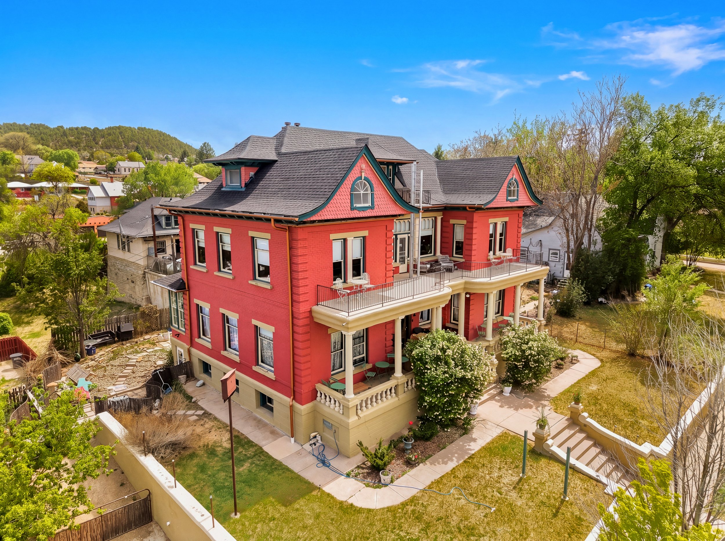 A large Victorian-style house painted red with beige accents, featuring a wrap-around porch, ornate roof design, and multiple levels. The yard has trees, potted plants, and concrete walkways, with neighboring houses and a hillside in the background under a clear blue sky.