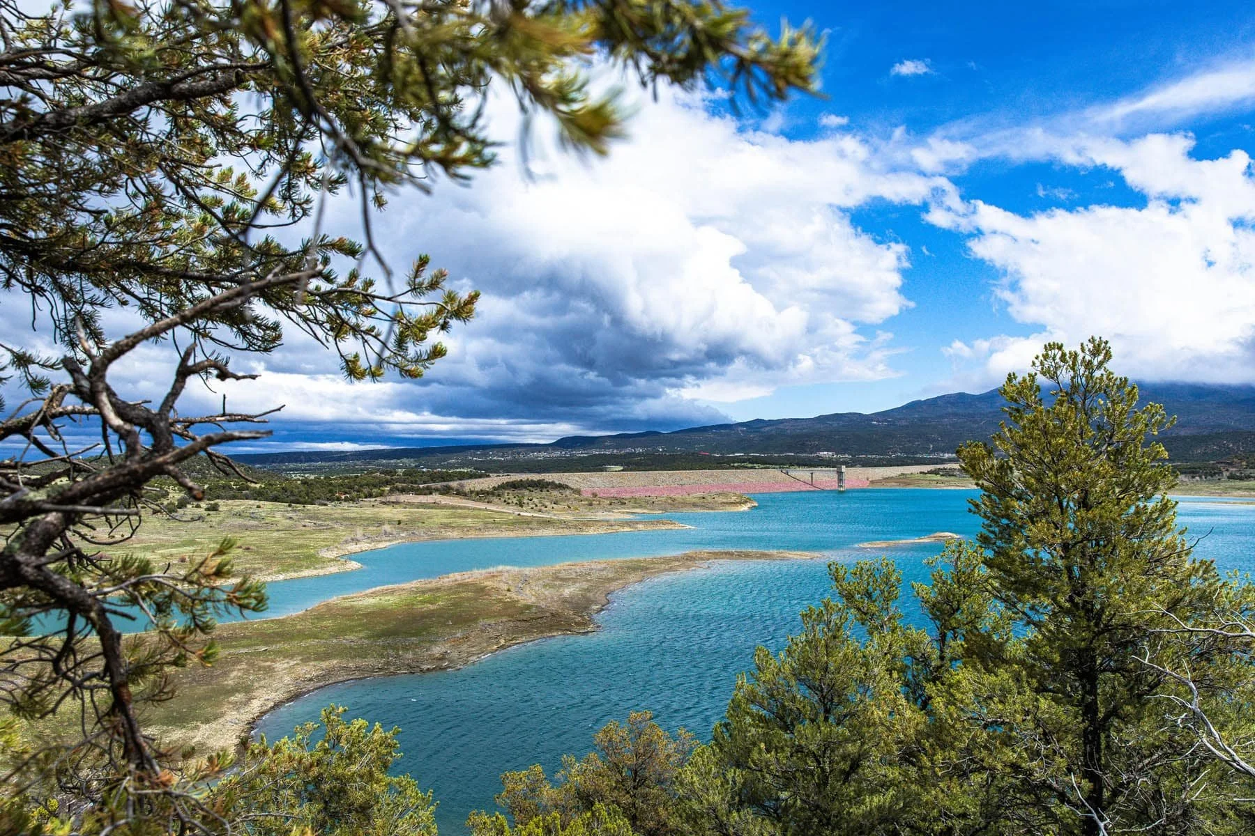 View of a large reservoir with blue water, surrounded by green trees and hills under a partly cloudy sky.