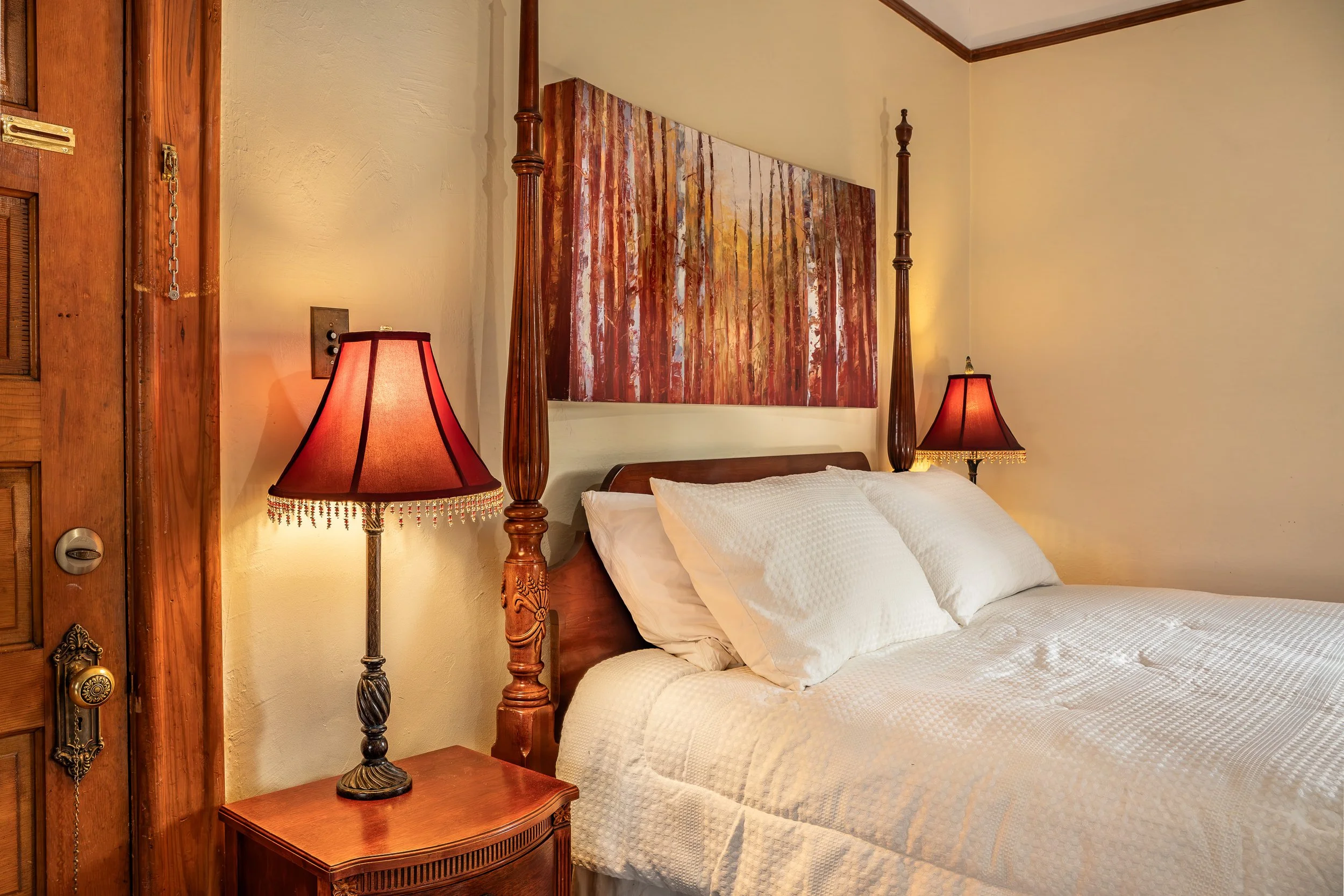 A bedroom with a wooden four-poster bed, white bedding, two matching table lamps with red shades, a colorful forest-themed painting above the bed, and a wooden nightstand.