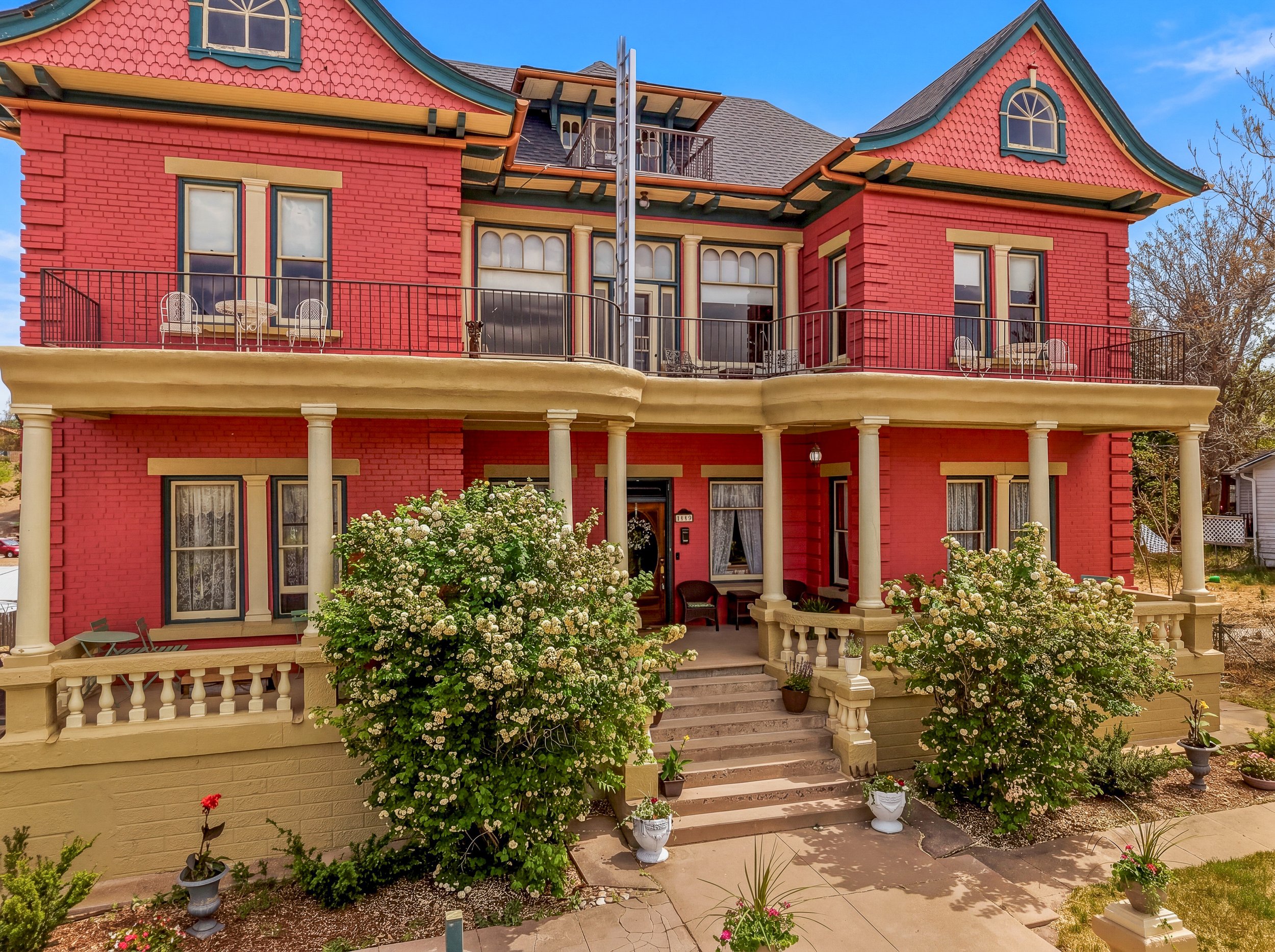 A large, red, Victorian-style house with detailed trim, multiple windows, and a balcony. A staircase leads to the front porch decorated with potted plants and flowers.
