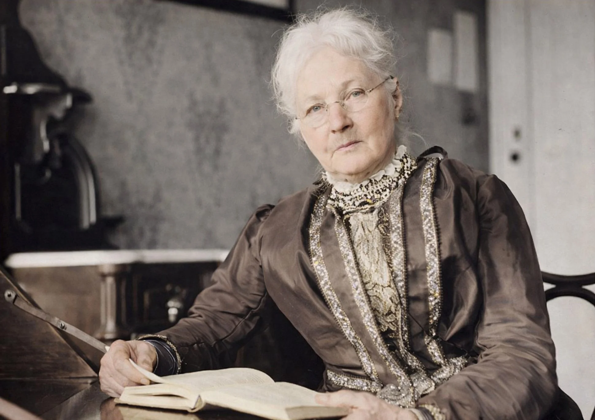 An elderly woman with white hair, glasses, and a stern expression, sitting at a desk with an open book and a pen, in a room with a gray wall.