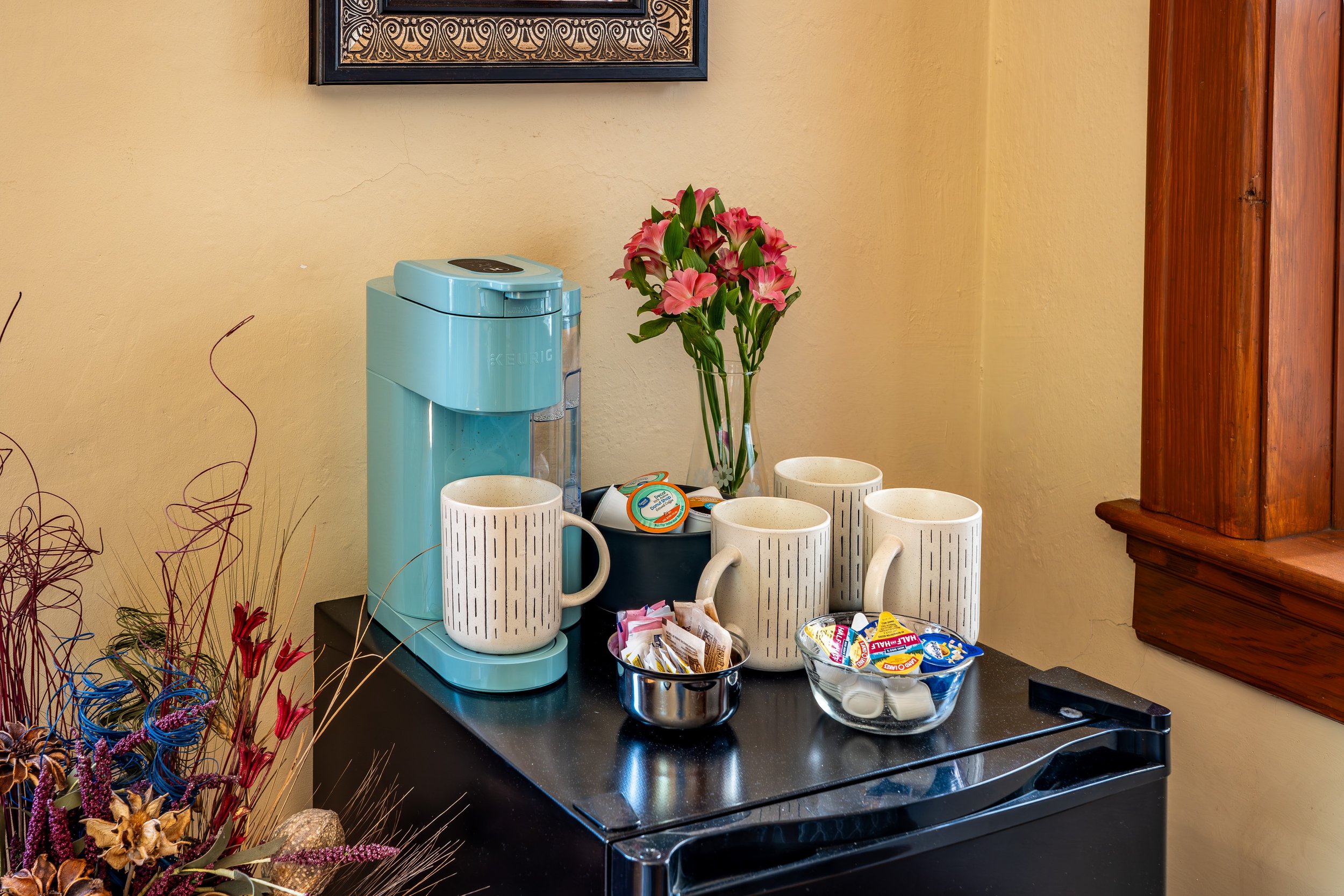 A kitchen or breakfast bar area with a Keurig coffee maker, four white mugs, a vase of pink lilies, and bowls of single-serving snacks on a black countertop. A framed decorative artwork hangs on a yellow wall, and part of a wooden cabinet is visible on the right side.