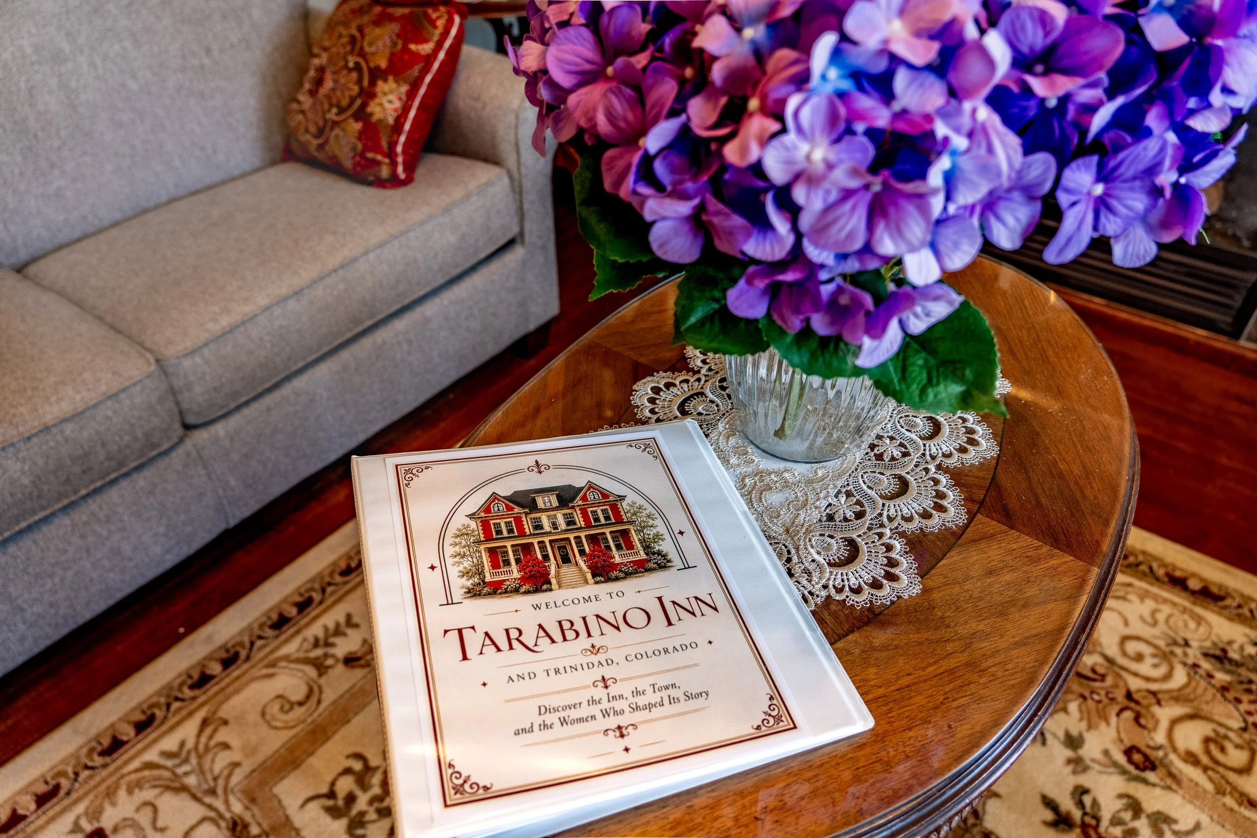 A wooden table with a floral lace doily, a brochure for Tarabino Inn, and a large purple hydrangea bouquet in a glass vase, with a beige sofa and ornate red pillow in the background.