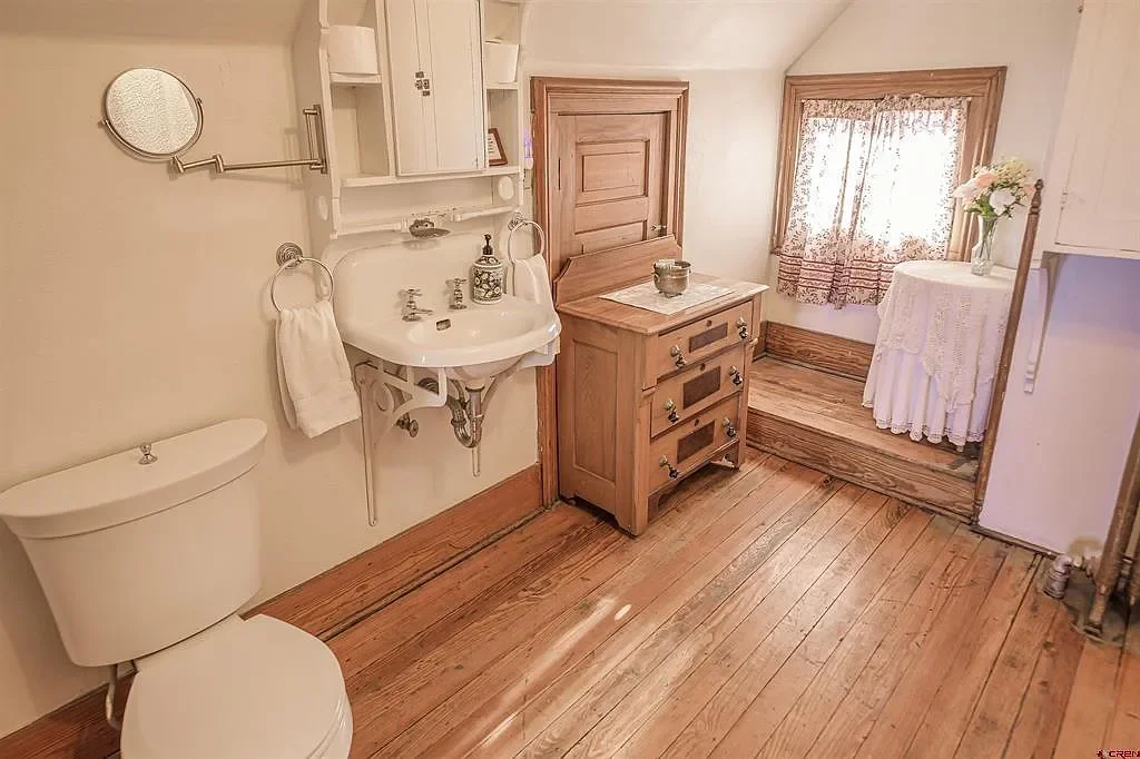 A vintage bathroom with a toilet, small white sink, and wooden furniture, including a chest of drawers and a small table with lace curtain in the window.