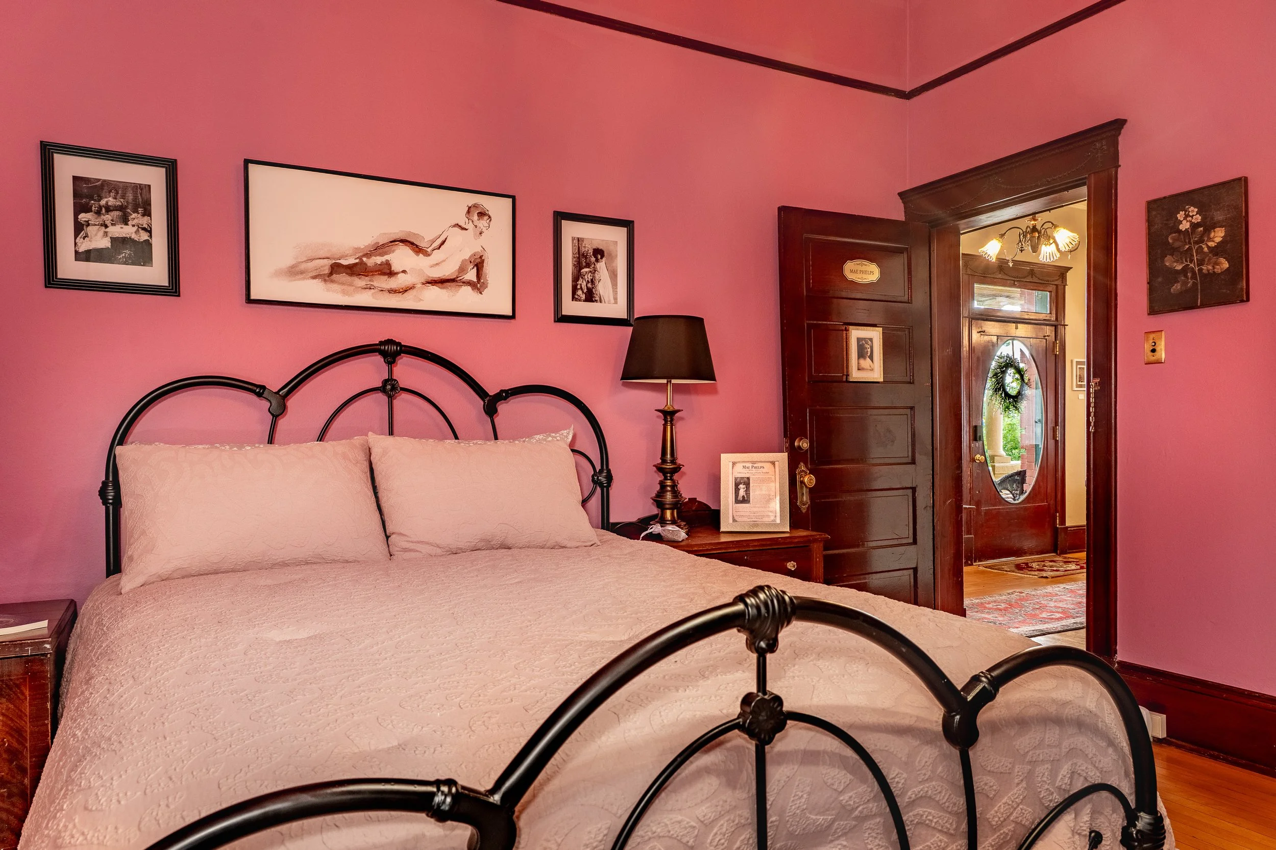 A bedroom with pink walls, a black metal bed frame, beige bedding, a wooden nightstand, a black lamp, framed artwork, and an open wooden door leading to a hallway with a wreath on the door and a chandelier.