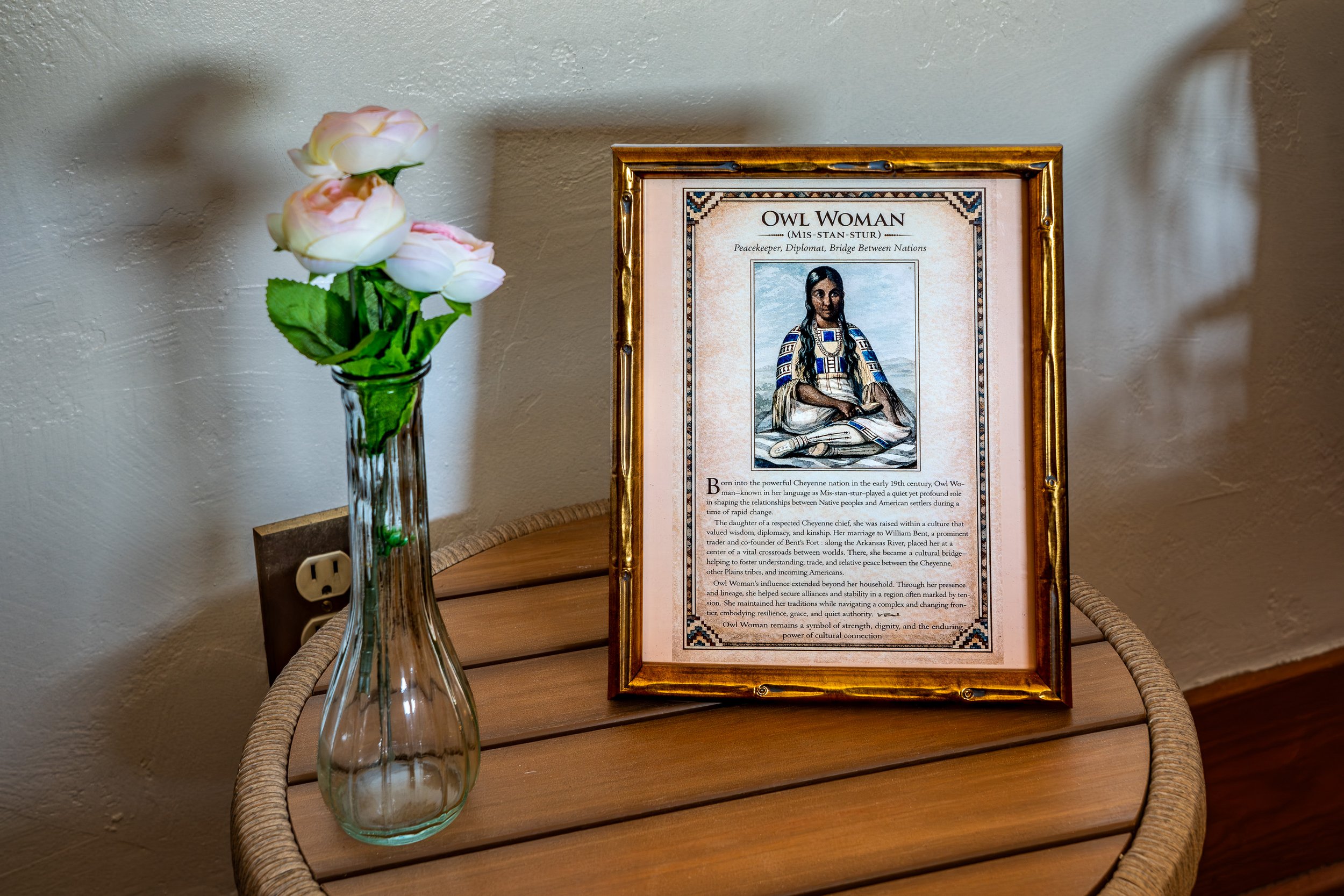 A wicker table holding a framed informational poster about Owl Woman, and a tall glass vase with white and pink artificial roses, placed near a wall with an electrical outlet.