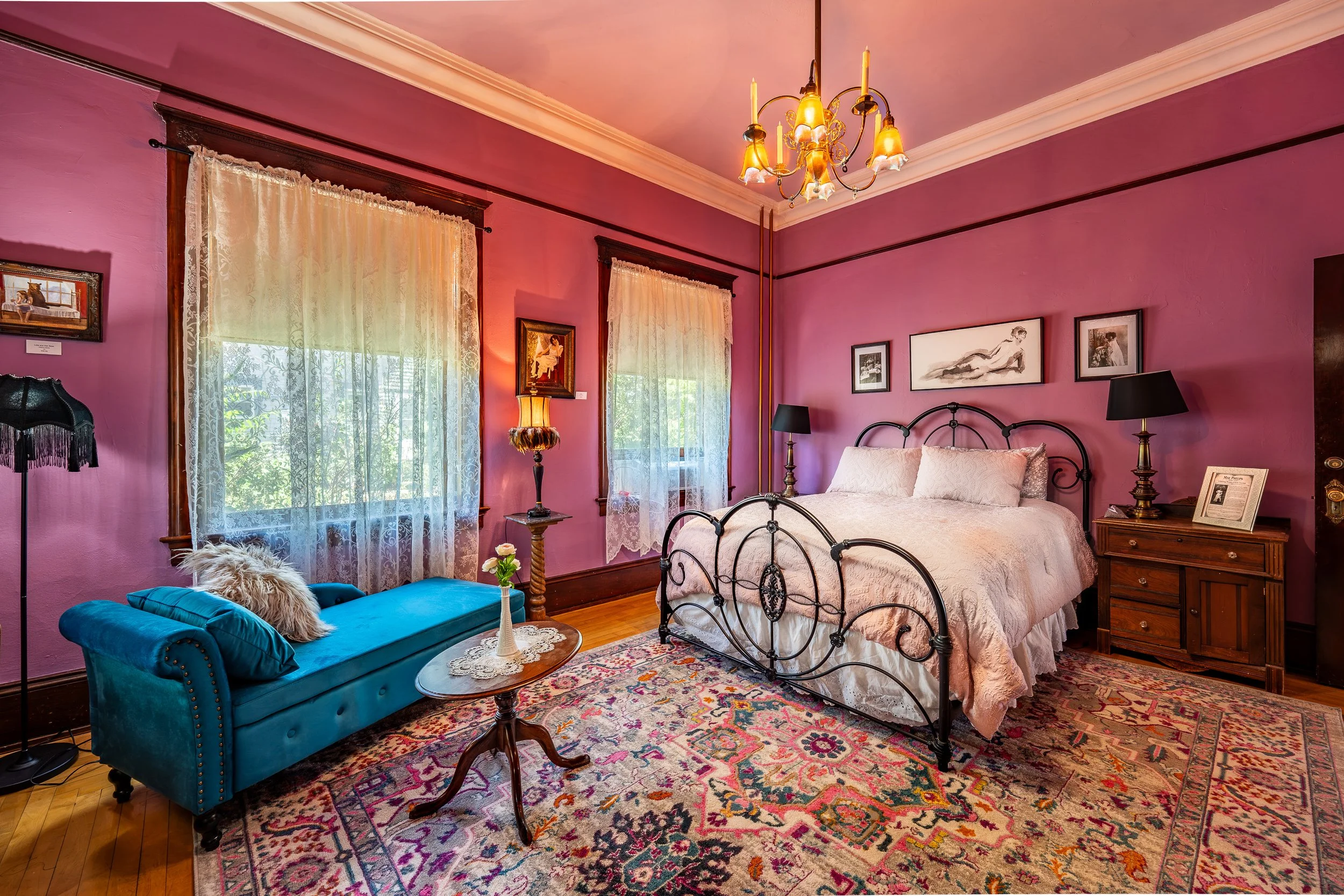 A vintage-style bedroom with purple walls, lace curtains, and hardwood floors. There is a black metal bed with white bedding, a wooden nightstand with a framed picture, a black lampshade, and a brass chandelier hanging from the ceiling. A turquoise tufted chaise lounge with a fur pillow, a small round table with a lace doily and flower arrangement, and a colorful patterned rug complete the decor.