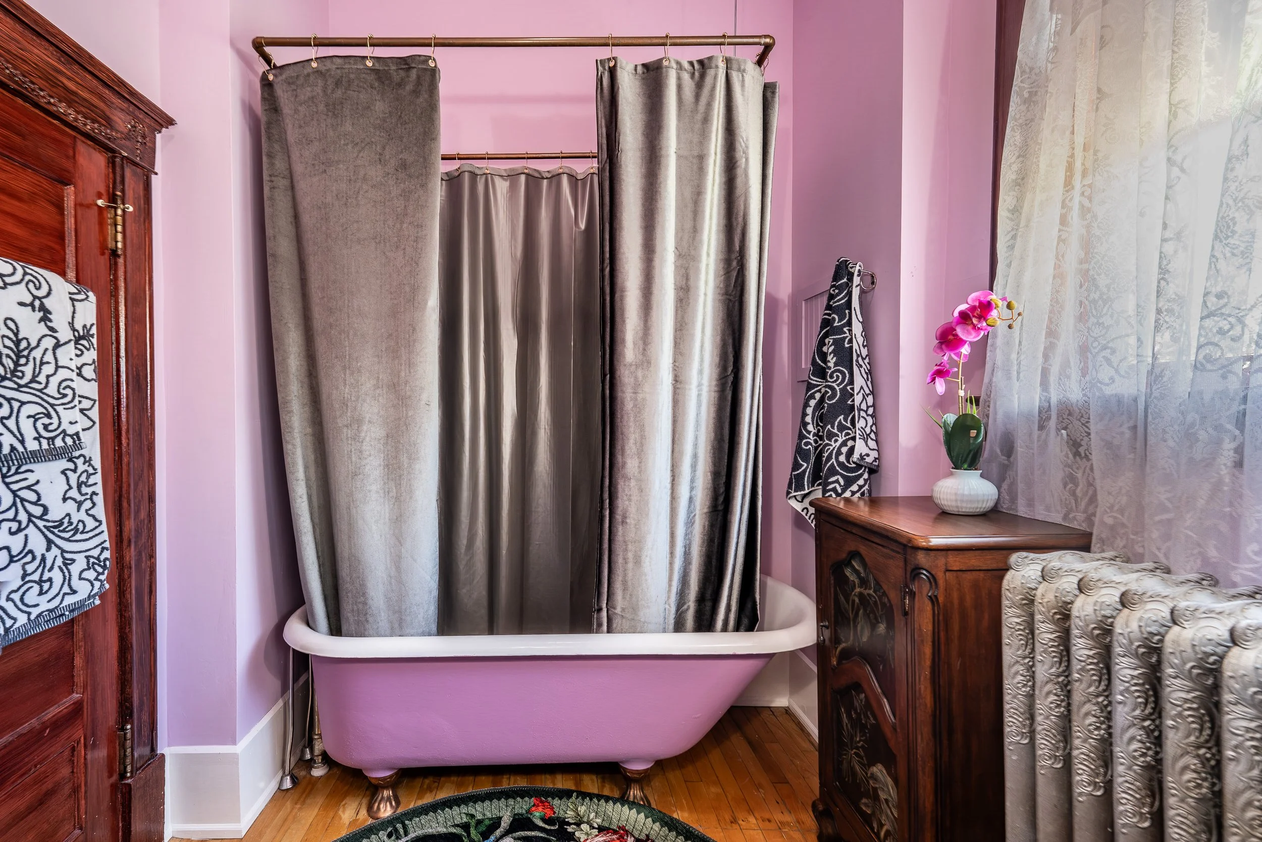 A vintage pink bathtub with a shower curtain, located in a bedroom with purple walls, a wooden dresser, an orchid plant, and lace curtains.
