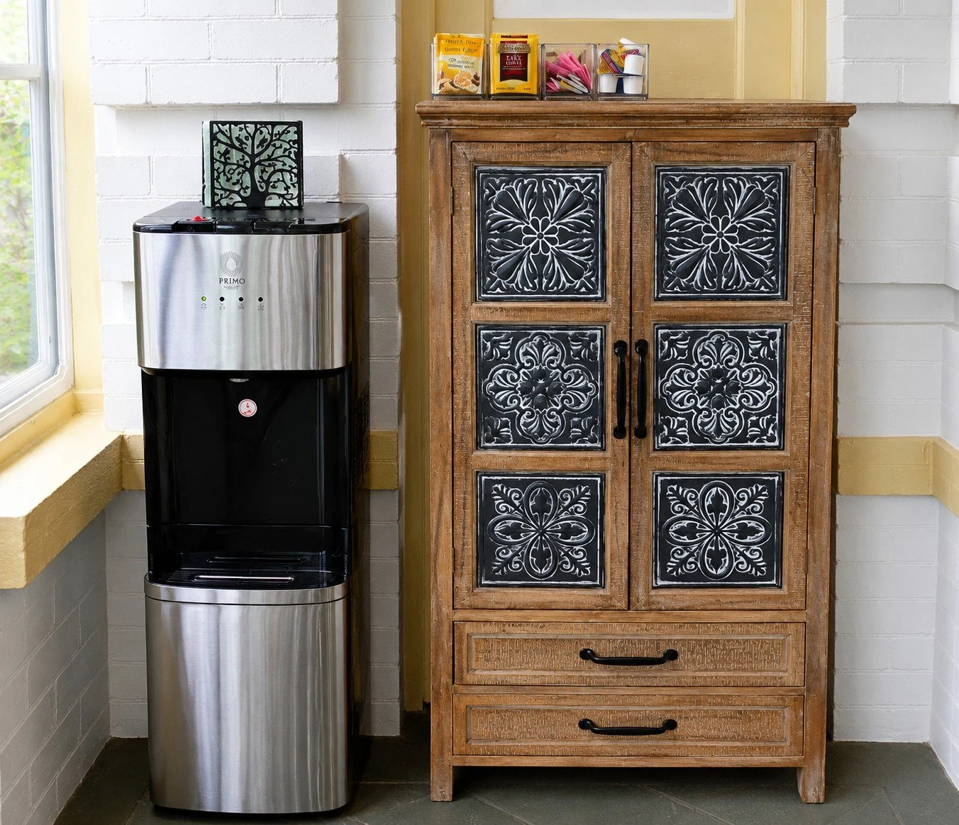 A water cooler next to a wooden cabinet with ornate black and white design on its doors, located near a window in a kitchen or break room.