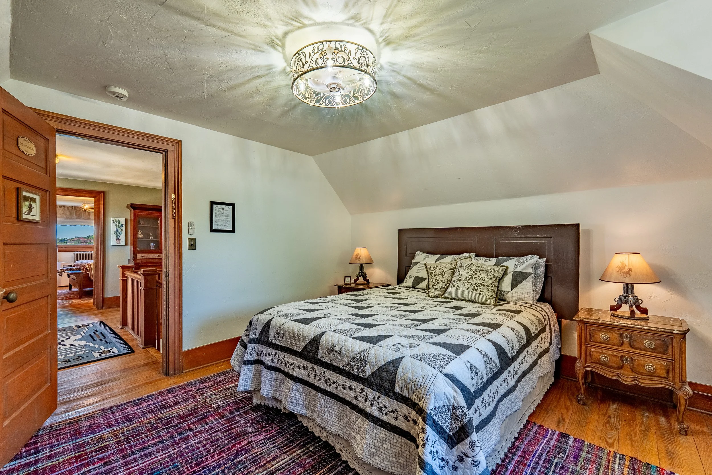 A bedroom featuring a bed with a black and white quilt, bedside tables with lamps, and decorative pillows. An ornate ceiling light casts shadows on the ceiling. The room has wood flooring and a colorful area rug. An adjacent room with a window and furniture is visible through an open doorway.