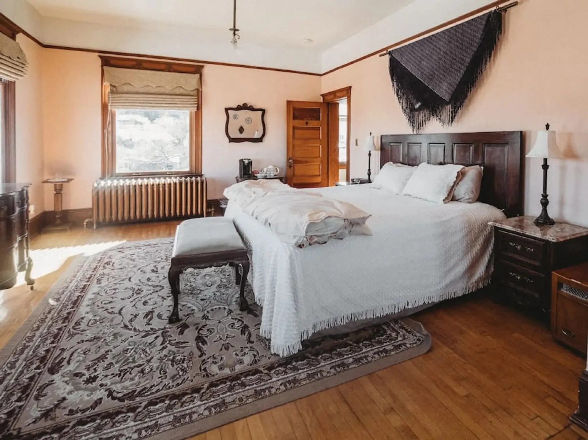 A cozy bedroom with a large bed covered in white linens and a fringed bedspread. The room features wooden trim, a patterned area rug, a window with a beige shade, and furniture including nightstands, a dresser, and a bench at the foot of the bed. Black wall hanging above the headboard and a rug on the floor.