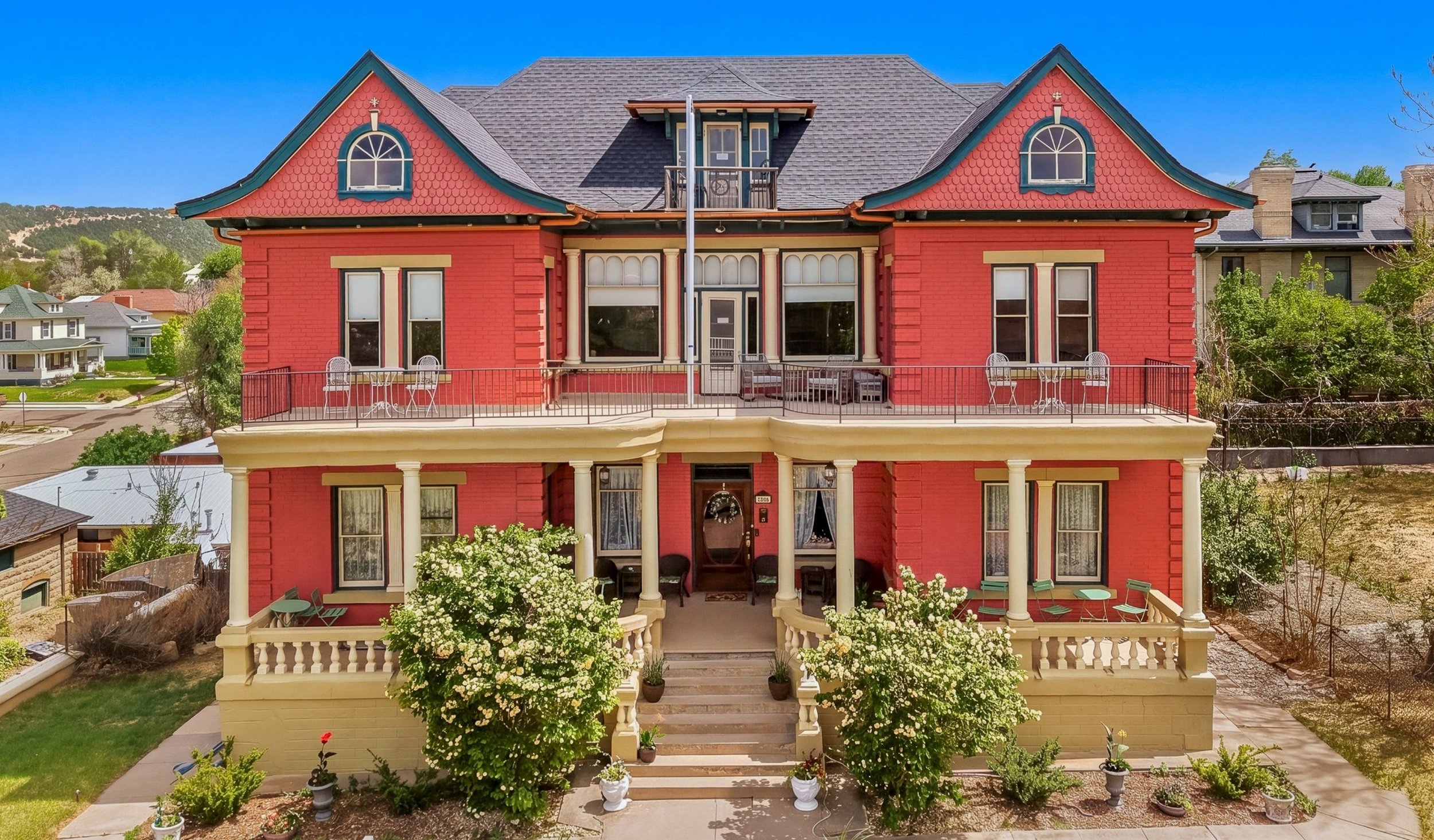 A large, colorful Victorian-style house painted red with cream accents and decorative trim. It has multiple stories, a prominent front porch, and a spacious balcony on the second floor. The house is surrounded by a well-maintained garden with blooming bushes and potted plants. Behind the house, there are neighboring homes and green hills under a clear blue sky.