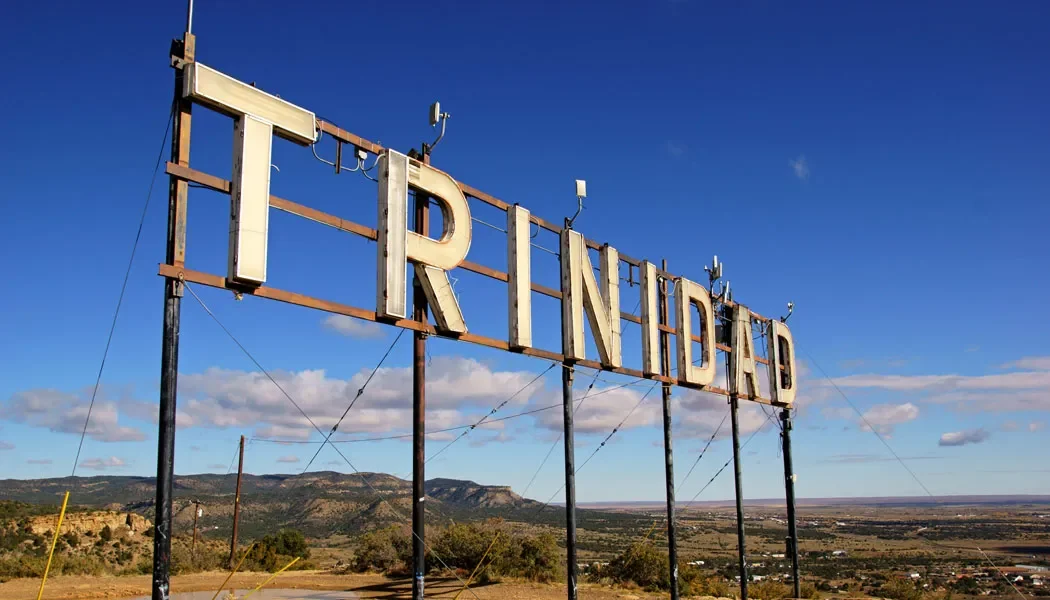 Large, weathered sign spelling 'TRINIDAD' on metal poles in a rural landscape with mountains in the background under a blue sky with clouds.