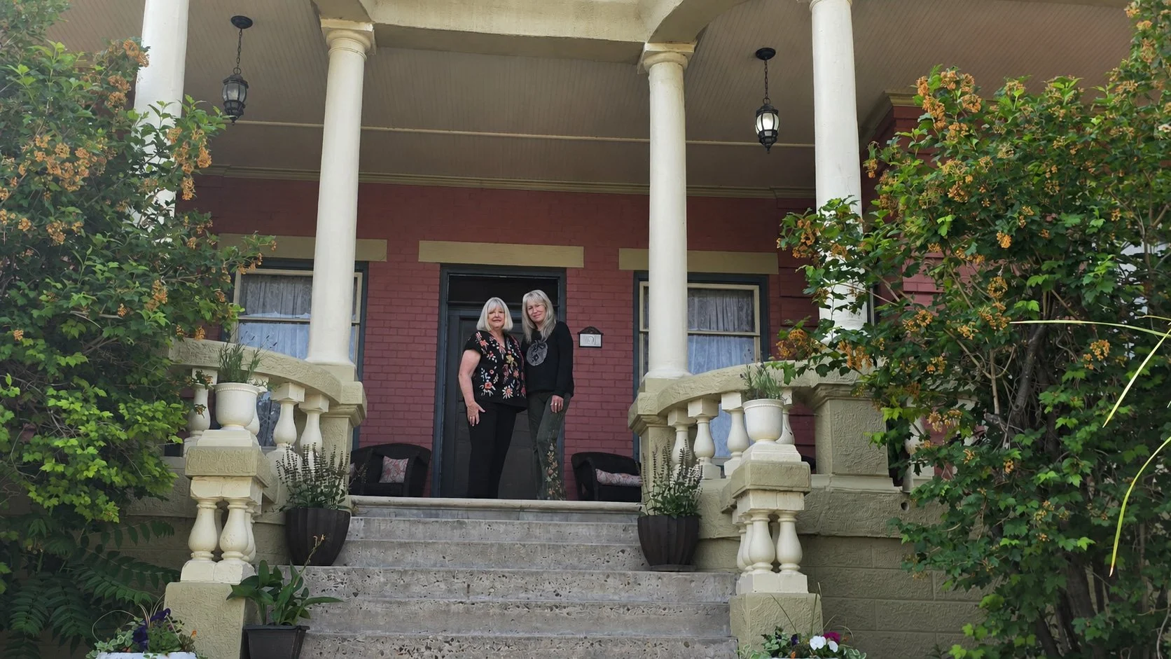 Two women standing on the front porch of a red brick house with cream-colored columns and railings, surrounded by green bushes and potted plants.