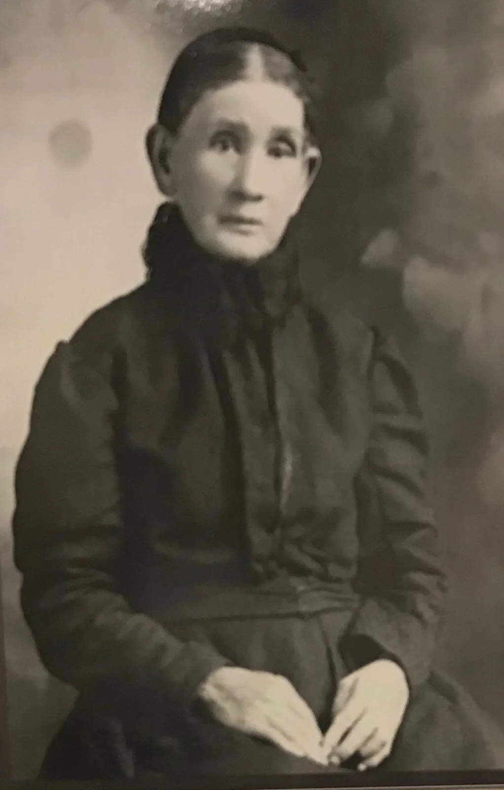A woman sitting with her hands in her lap, wearing a dark jacket, with her hair tied back, looking at the camera in a black and white photo.