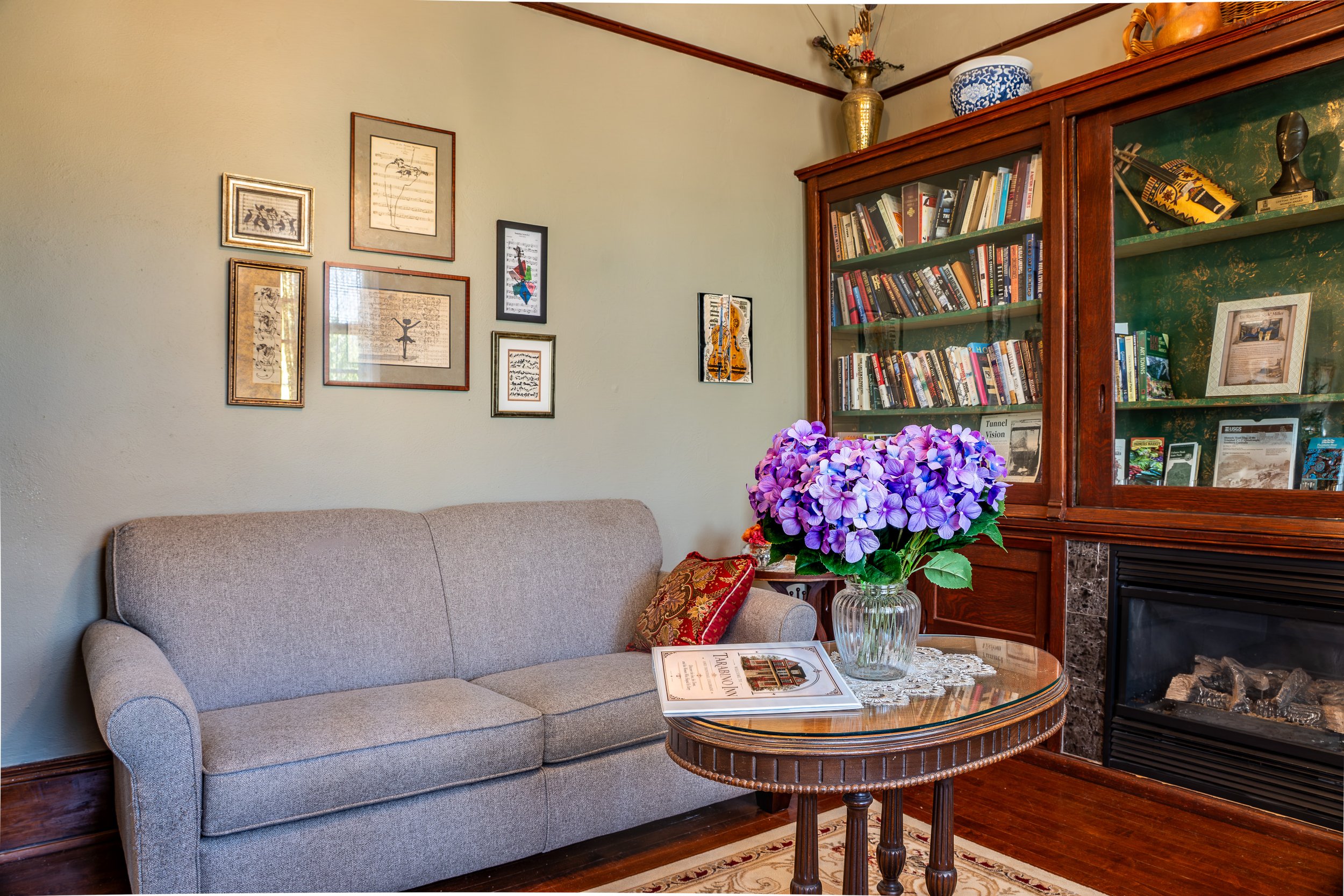 Living room with a beige sofa, a wooden coffee table with a book, a lace tablecloth, and a large glass vase with purple hydrangeas. The background features a framed wall art collection, a wooden bookshelf filled with books, a vase with flowers, and a fireplace on the lower right.