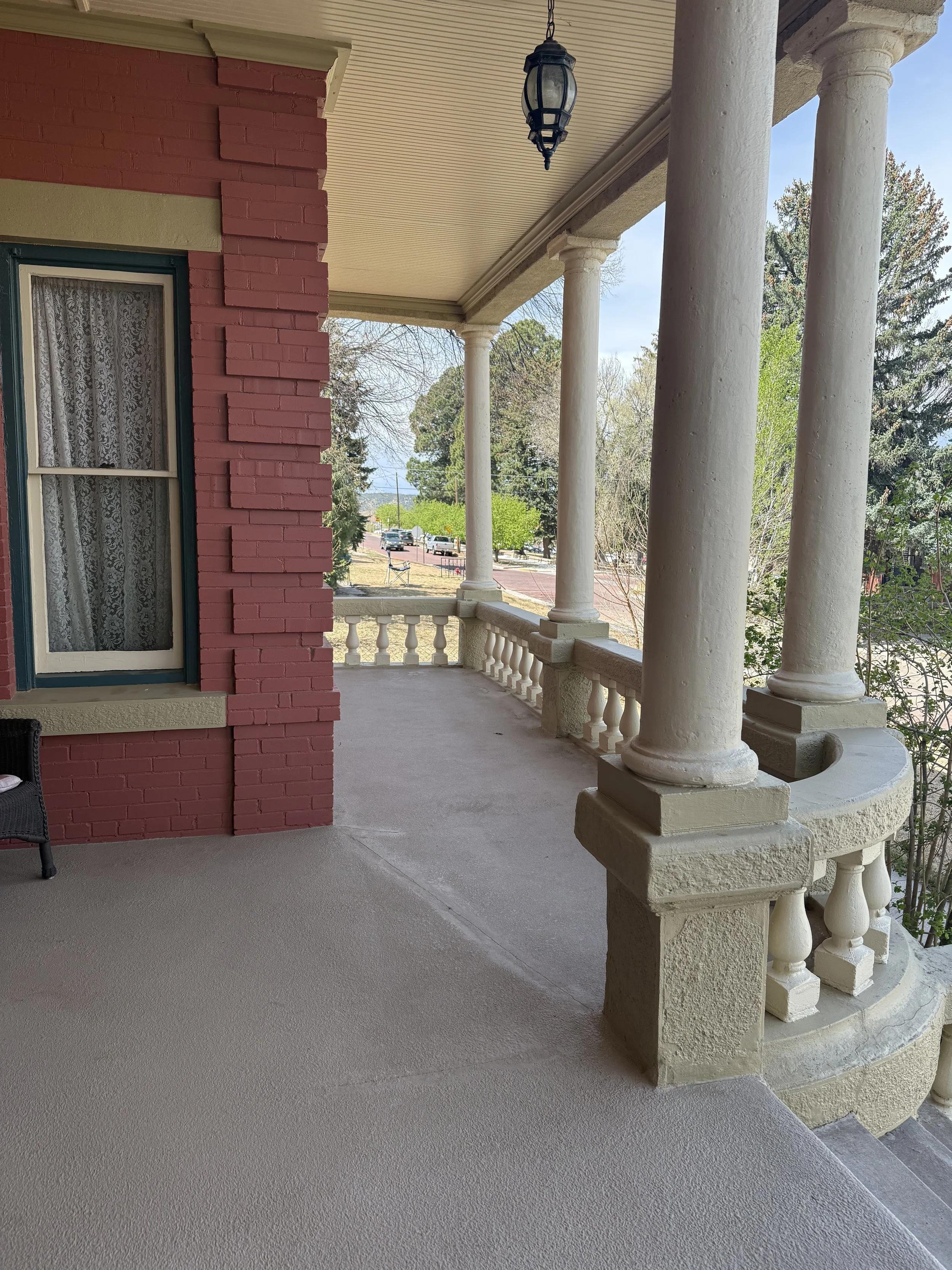 View of a porch with white columns, a brick wall with a window, and a textured concrete floor. Trees and a street with cars are visible in the background.