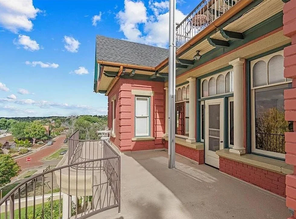 View of a rooftop balcony of a colorful Victorian-style building with large windows, a door, a railing, and a small outdoor chair, overlooking a quiet neighborhood with trees and houses under a partly cloudy sky.