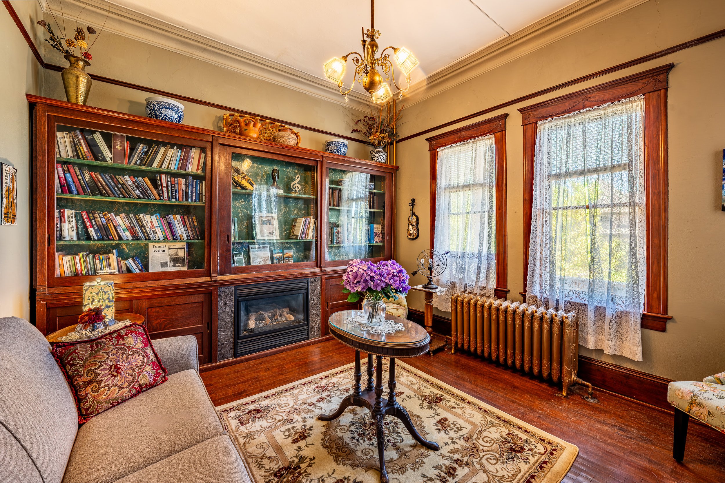 Living room with a large wooden bookshelf, a sofa with decorative pillows, a small round table with a vase of purple flowers, and two windows with lace curtains. The room has a wooden floor, a floral rug, and a brass radiator.