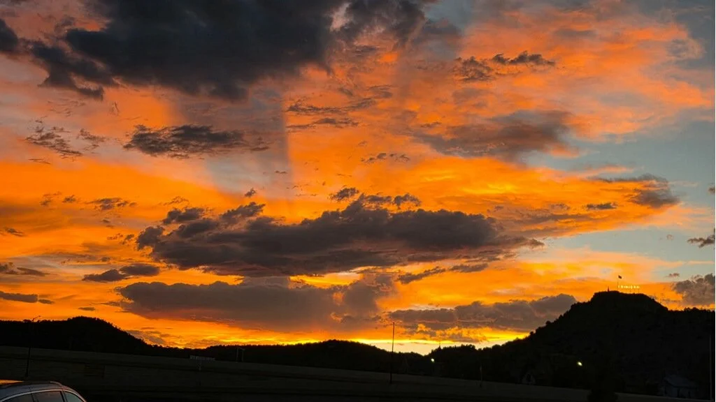 Sunset sky with orange and yellow clouds over silhouette hills, with a few lights visible at the bottom.