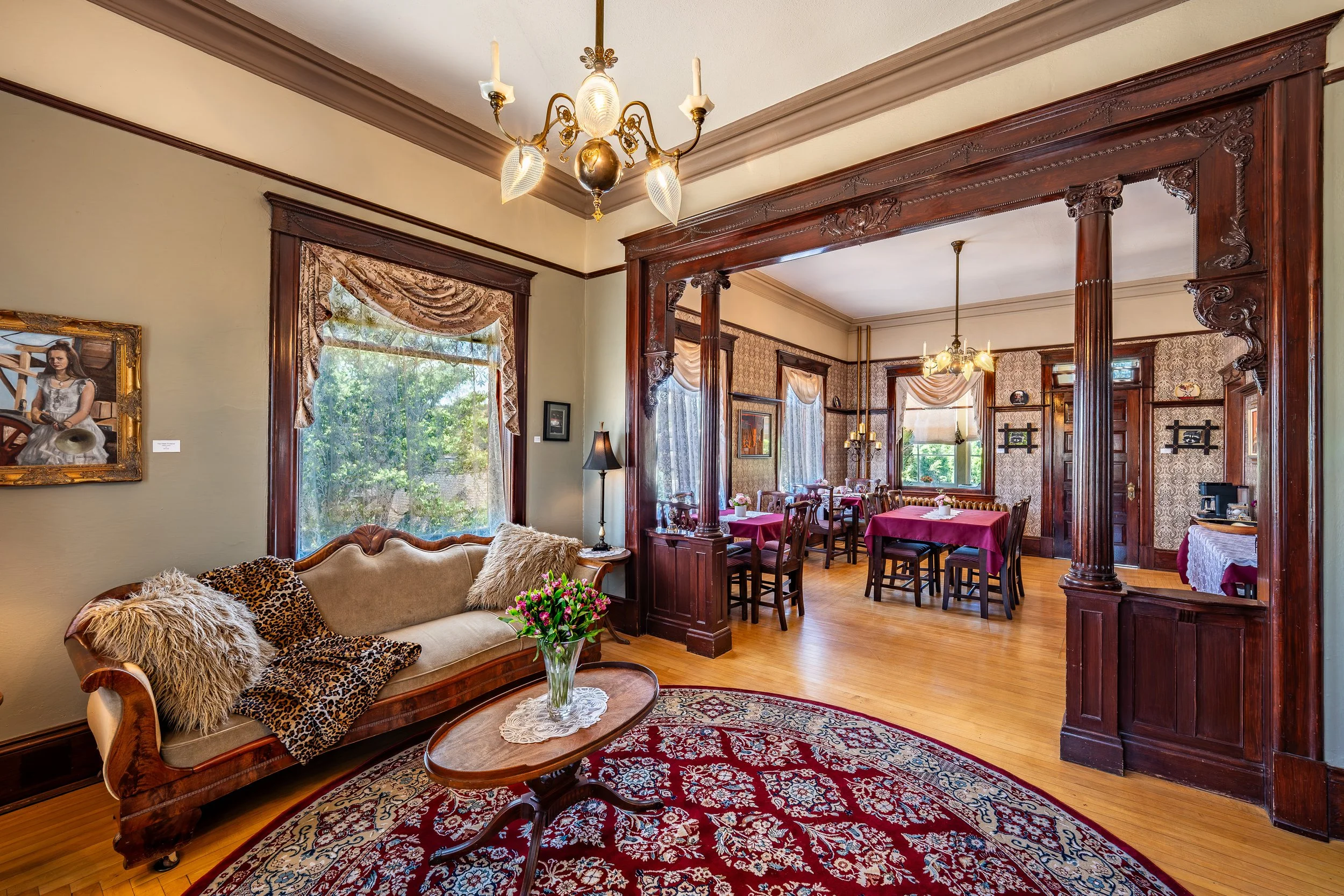 A Victorian-style living room with a cream-colored sofa, decorated with fur and leopard print pillows, a wrought iron coffee table with a floral arrangement, and large windows with lace and fabric curtains. The room is open to a dining area with dark wood furniture, patterned wallpaper, and wood trim, featuring a chandelier and pendant lights. The floor is hardwood and an ornate rug is placed under the coffee table.