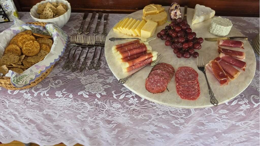 Charcuterie board with slices of cured meats, cheese, and grapes, alongside a basket of crackers on a lace tablecloth.