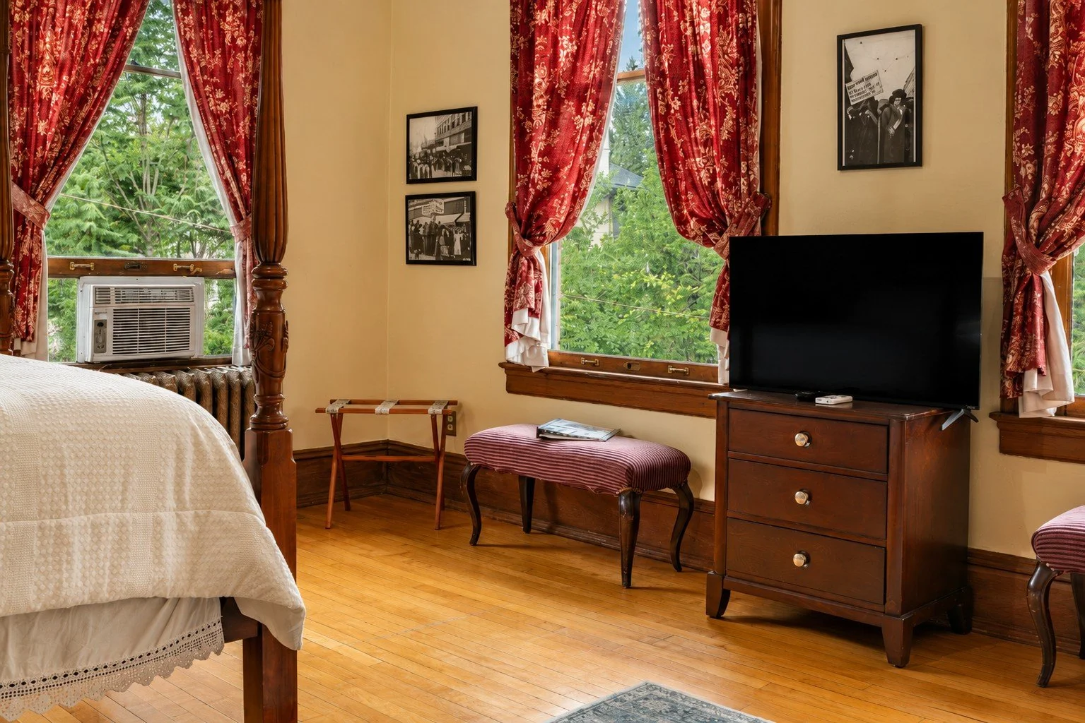 A cozy bedroom with a large wooden canopy bed, red and floral curtains, a window air conditioning unit, vintage framed black-and-white photos, a wooden dresser with a flat-screen TV, a small striped bench, a magazine, and hardwood floors.