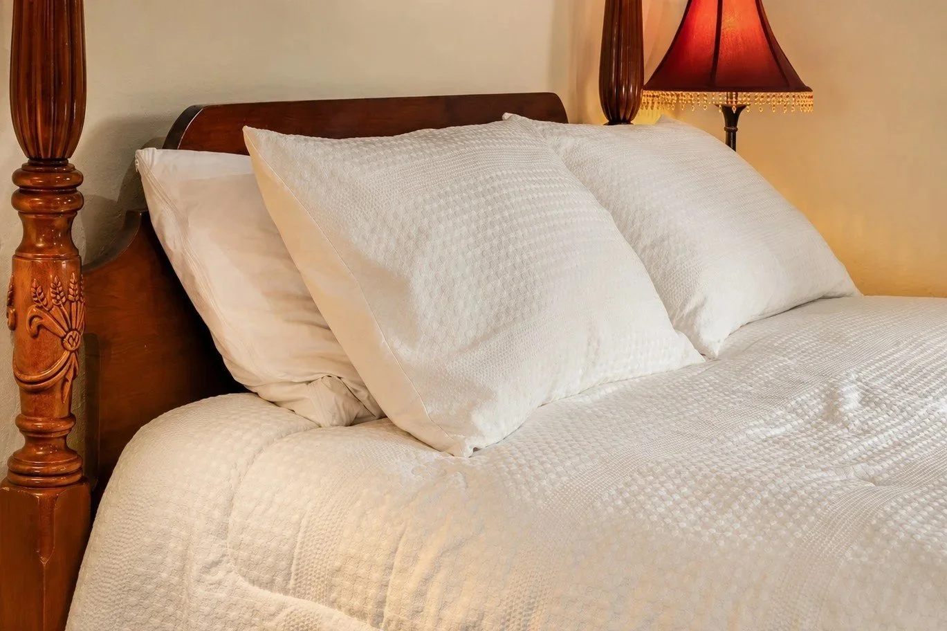 A neatly made bed with white textured bedding and large white pillows, wooden bed frame, and a bedside lamp with a red shade.