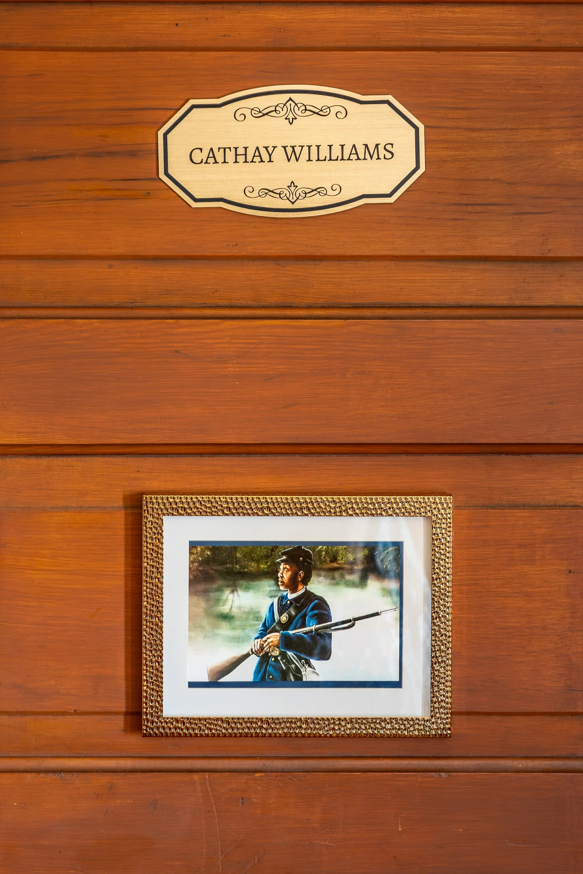 A wooden wall with a wall plaque that says 'Cathay Williams' and a framed portrait of a woman in a military uniform holding a rifle.