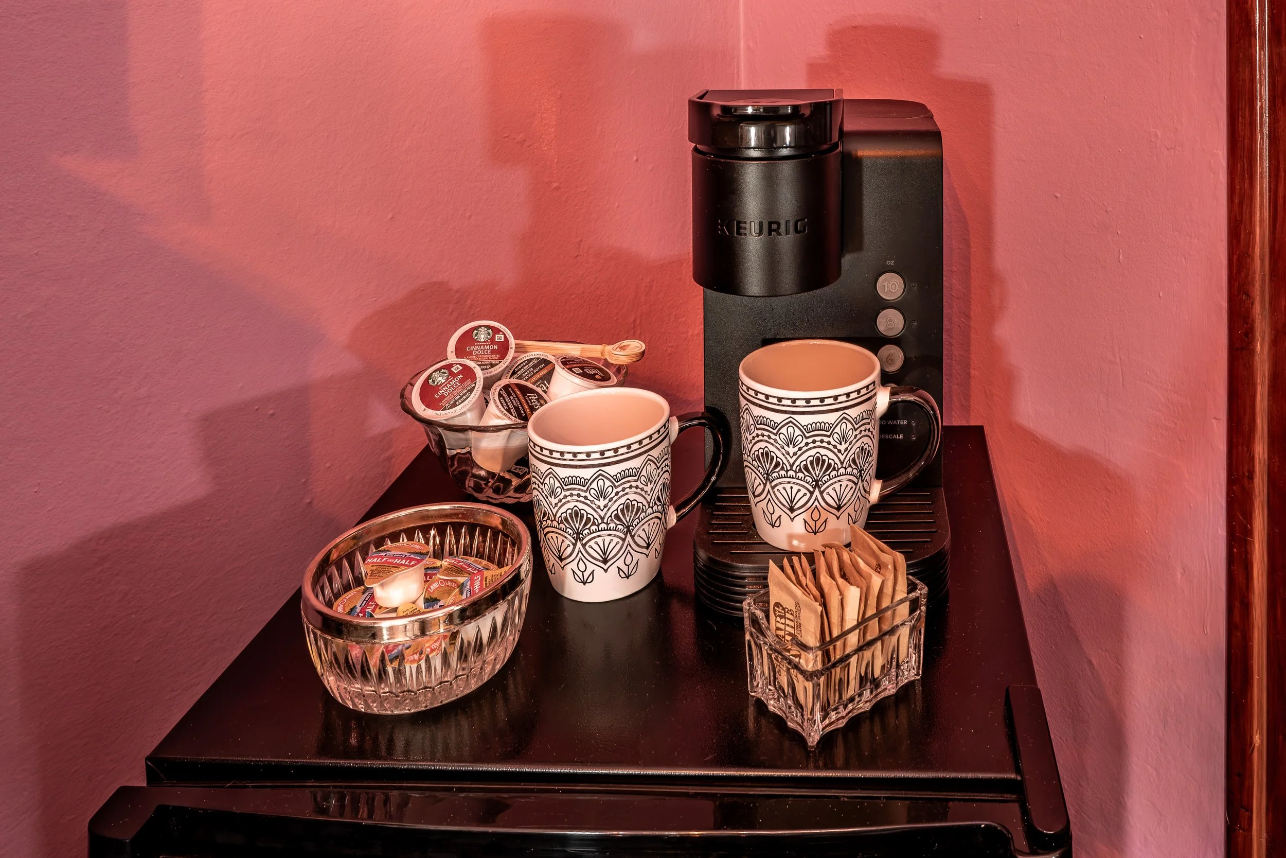 A coffee station with a Keurig coffee maker, two decorative mugs, a glass bowl filled with half & half coffee creamer cups, a glass container holding sugar packets, and some brown paper sugar bags on a black counter against a pink wall.