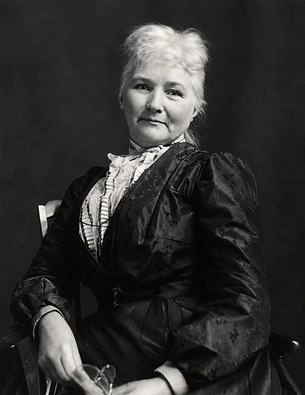 Black-and-white portrait of an older woman with short, tousled white hair, wearing Victorian-style clothing, seated on a chair, with a dark plain background.