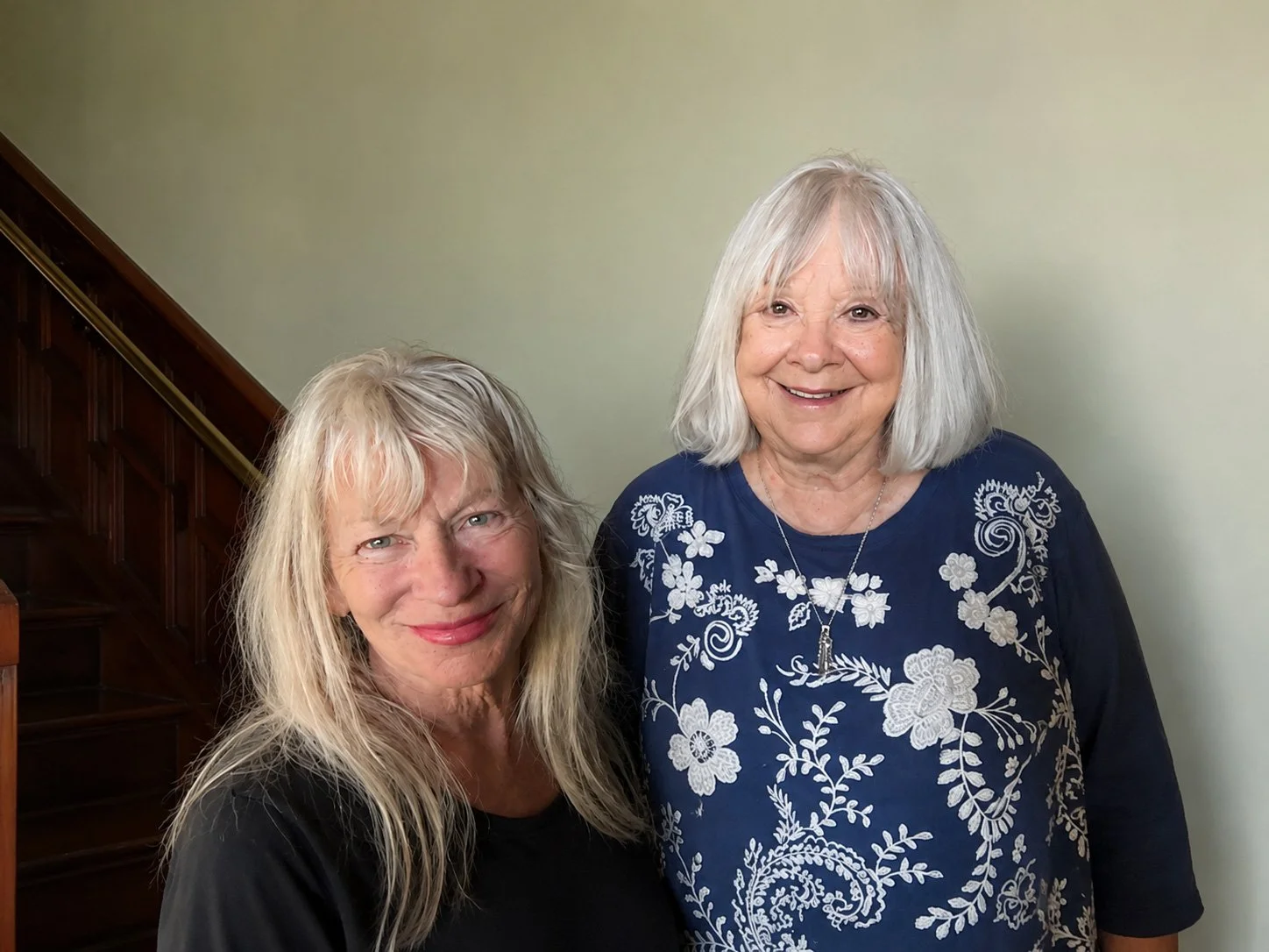 Two elderly women smiling indoors, one with long blonde hair and the other with short white hair, standing near a wooden staircase, against a pale yellow wall.