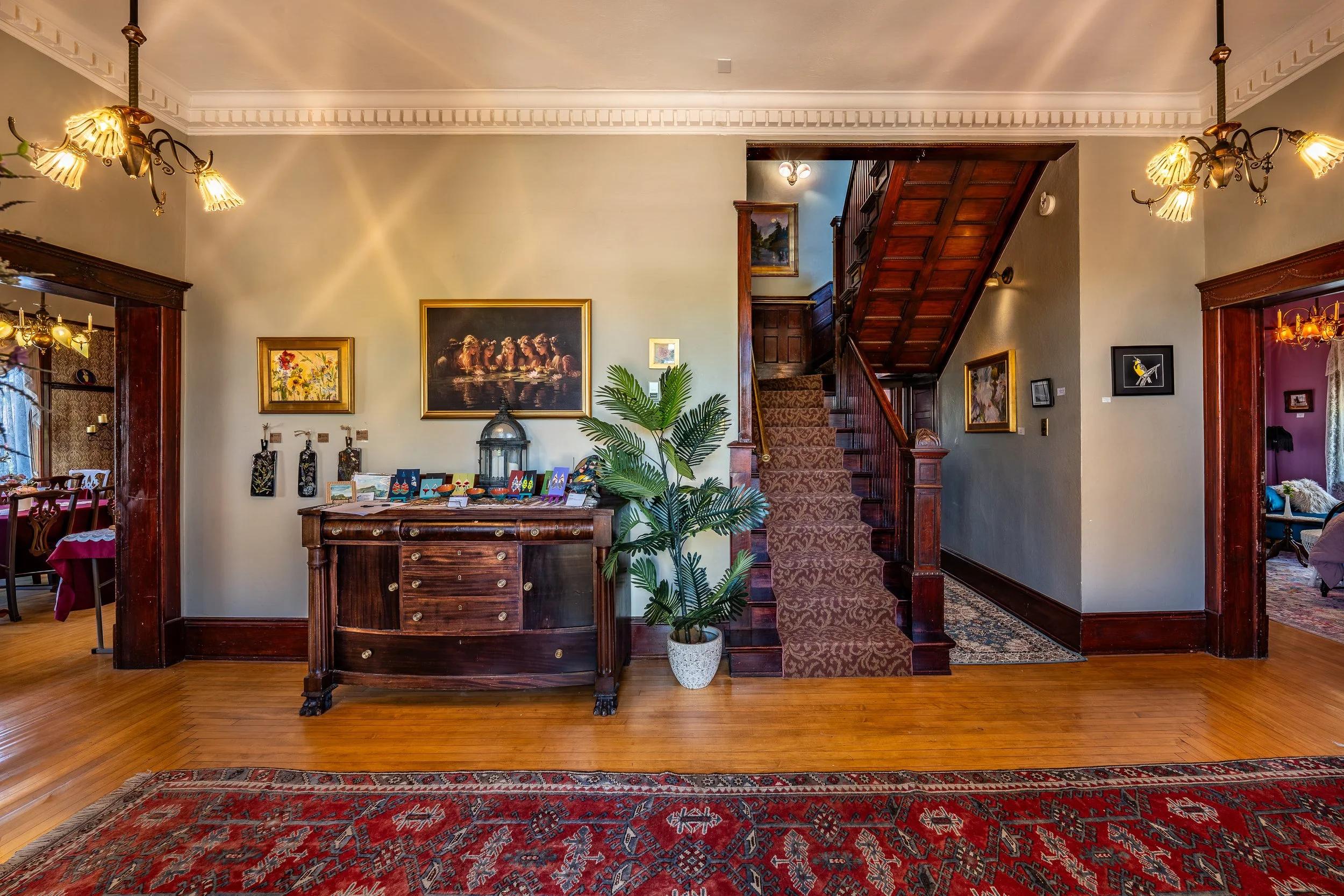 Interior view of a home with wooden floors, a decorative area rug, a wooden cabinet, a staircase with a patterned carpet, and framed artwork on the walls.