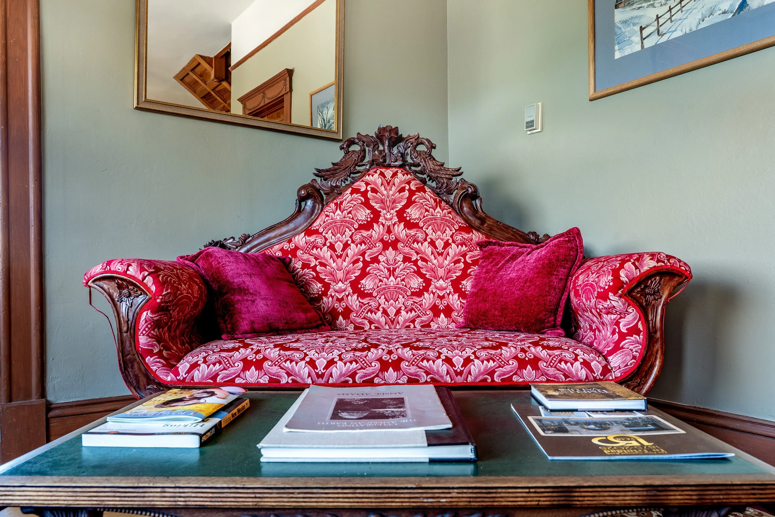 A wooden carved antique sofa with red and pink patterned upholstery, placed against a pale green wall. There are framed pictures hanging on the wall, including a landscape painting with a snow scene.