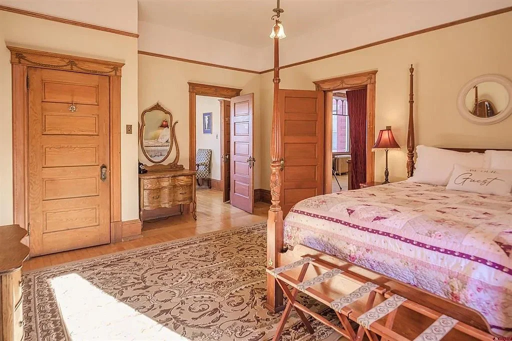 A bedroom with wooden furniture including a four-poster bed, a dresser with a mirror, and a wardrobe. There is a bed with a quilt and pillows, a lamp on the nightstand, and an ornate rug on the hardwood floor. Two open doors reveal a hallway and another room with curtains and a chair.
