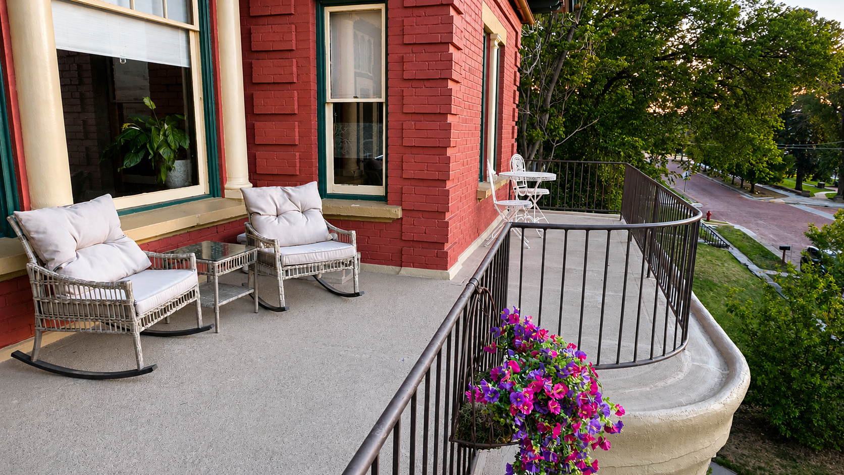 A front porch or balcony with two white wicker armchairs with cushions, a glass-topped table, a small white metal table with two chairs, and a potted plant with pink and purple flowers, overlooking a street with trees and houses.