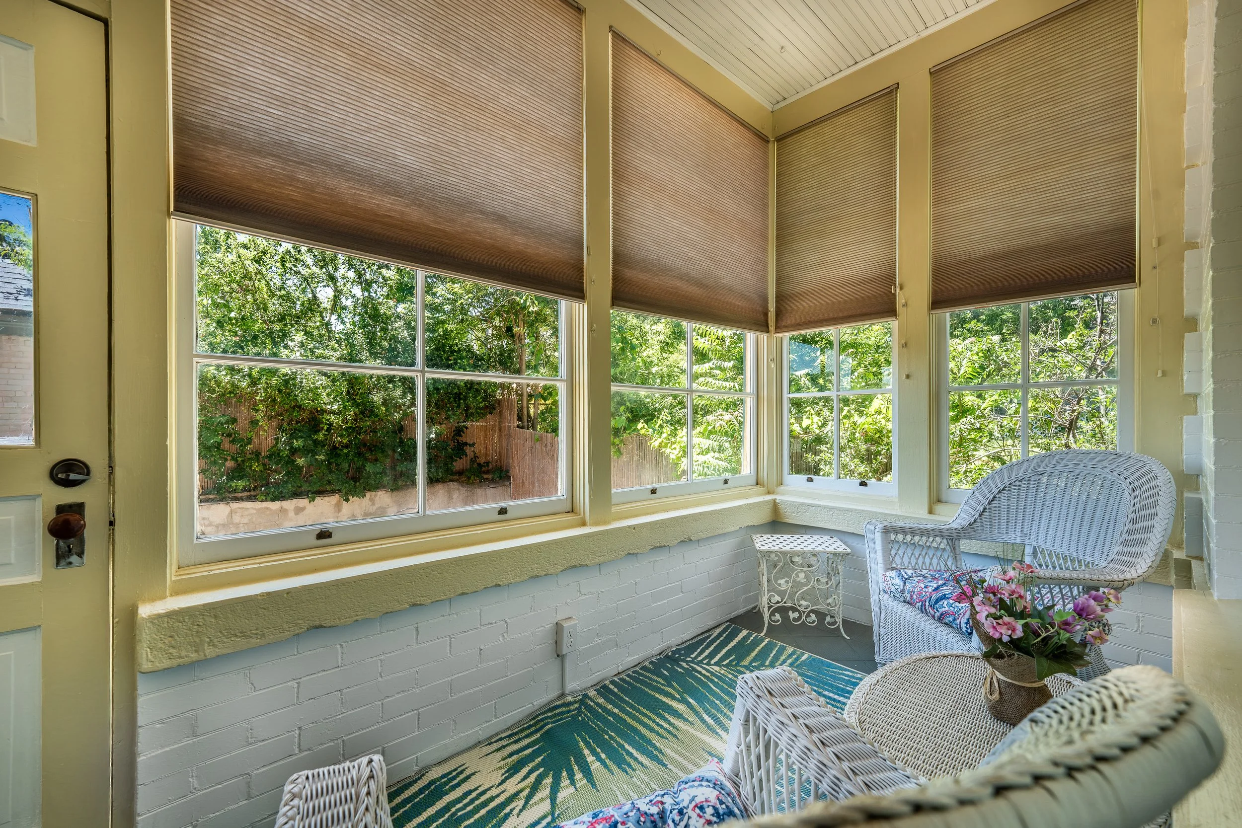 A cozy sunroom with large windows and wooden blinds, wicker chairs with floral cushions, a small table, and potted flowers, overlooking a green garden.