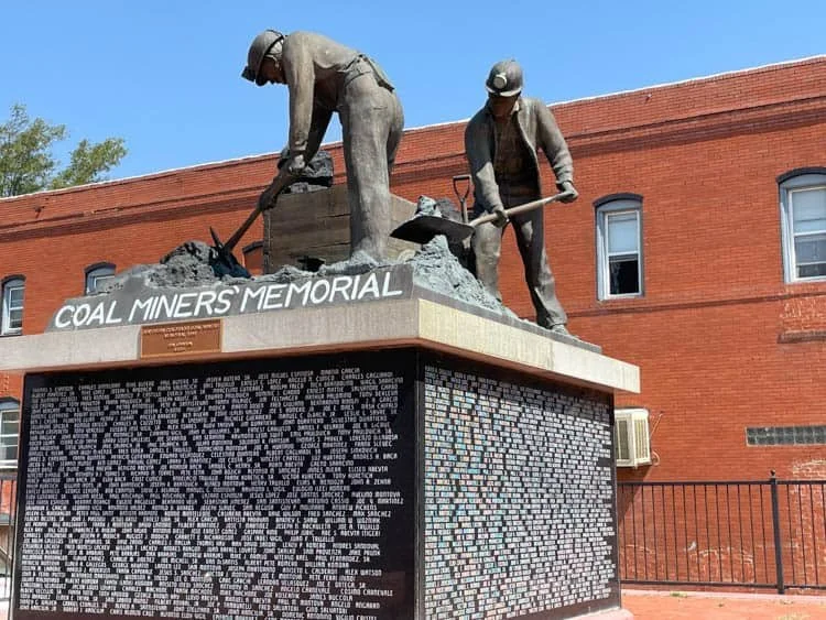 A memorial monument dedicated to coal miners features statues of two miners working, with a brick building in the background and a clear blue sky.