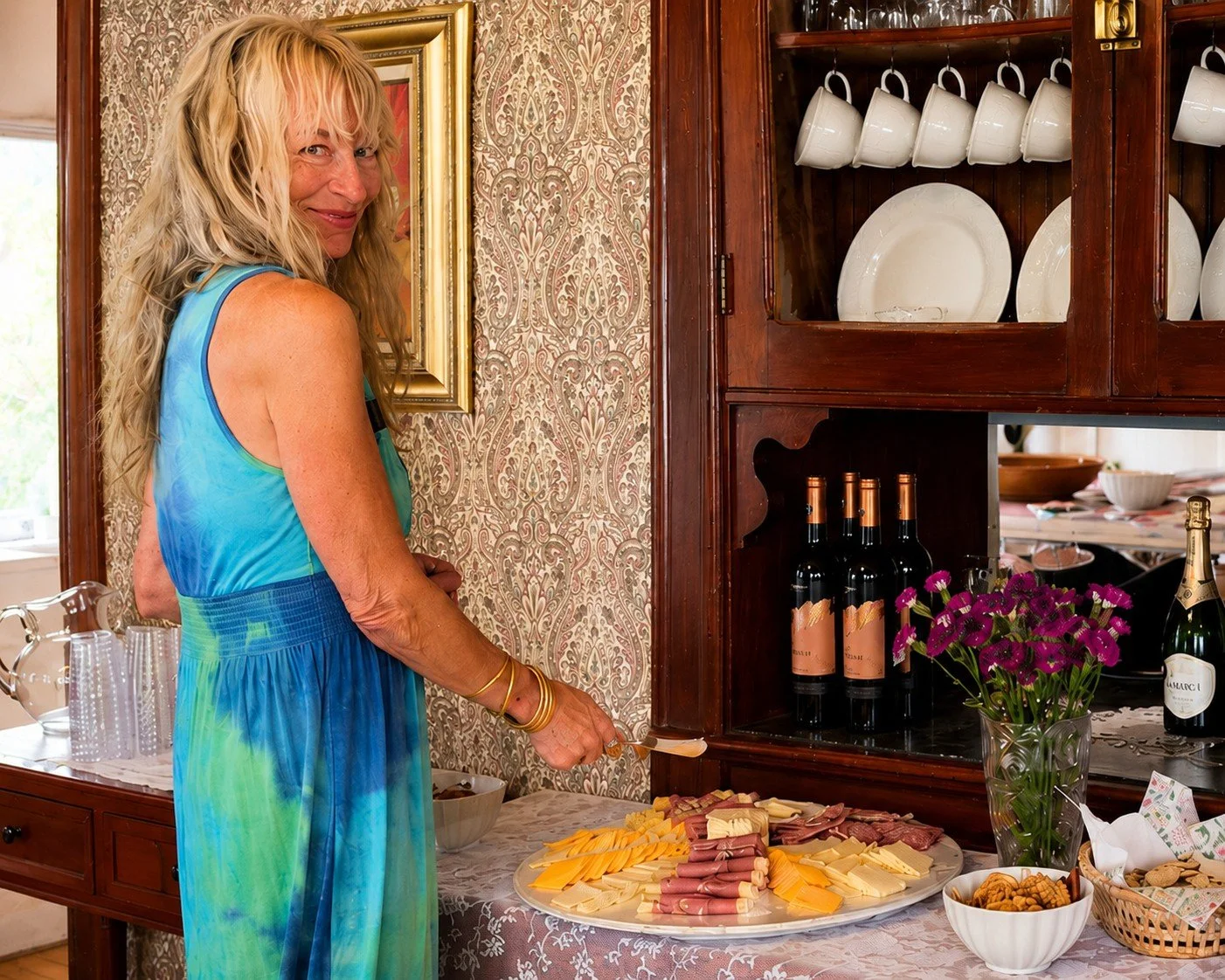 A woman with blonde hair in a blue and green dress stands next to a table with cheese, meats, and snacks, in a room with a wooden cabinet and patterned wallpaper.